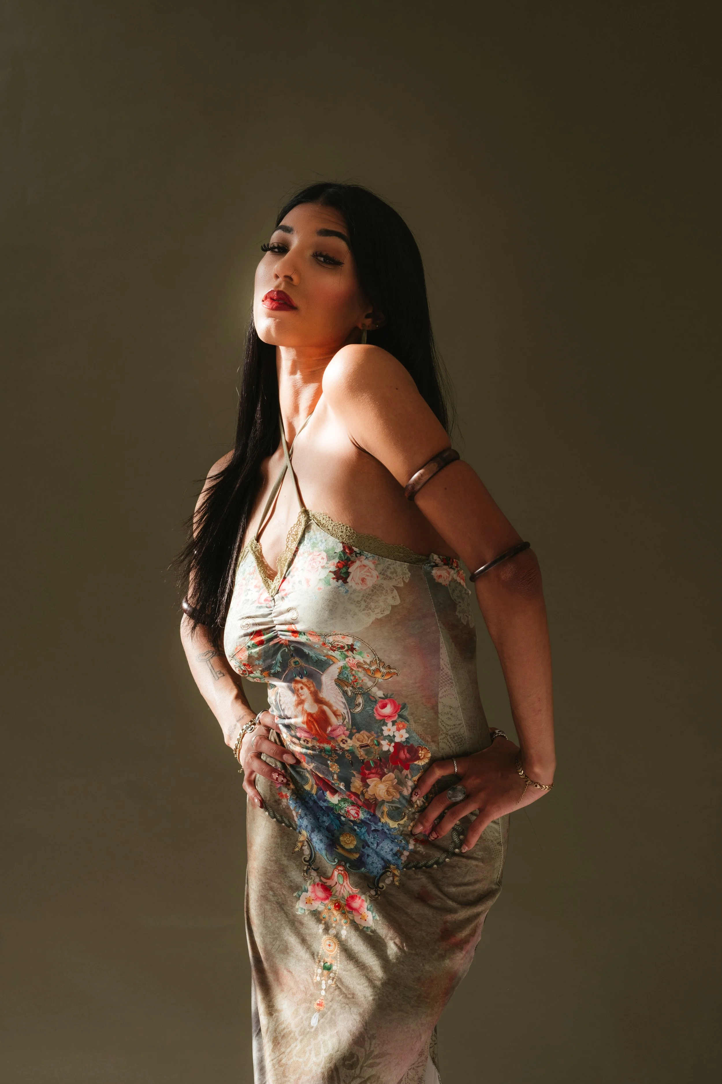A woman with long black hair and bold makeup, wearing a floral and embroidered dress, standing against a plain background.