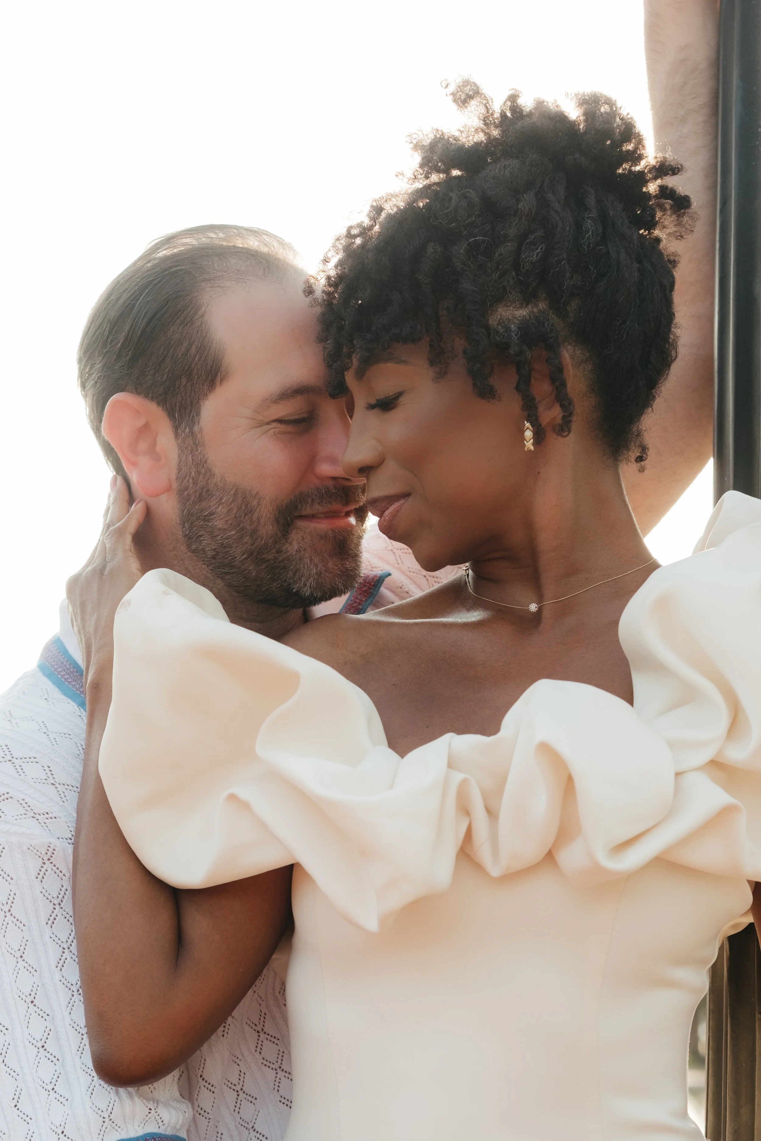 A couple embracing and about to kiss, with closed eyes, indoors with bright light behind them. The woman has short curly hair and is wearing a white dress with ruffled sleeves, and the man has dark hair and a beard, wearing a white shirt with a blue 