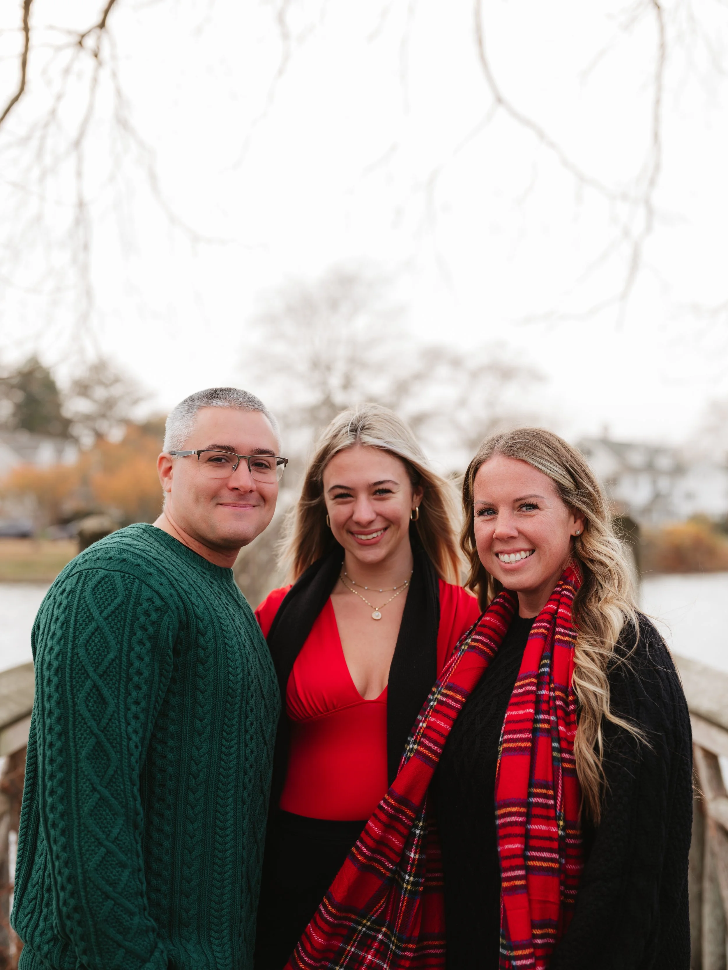 Three people smiling outdoors on a wooden bridge in autumn, with trees and houses in the background.