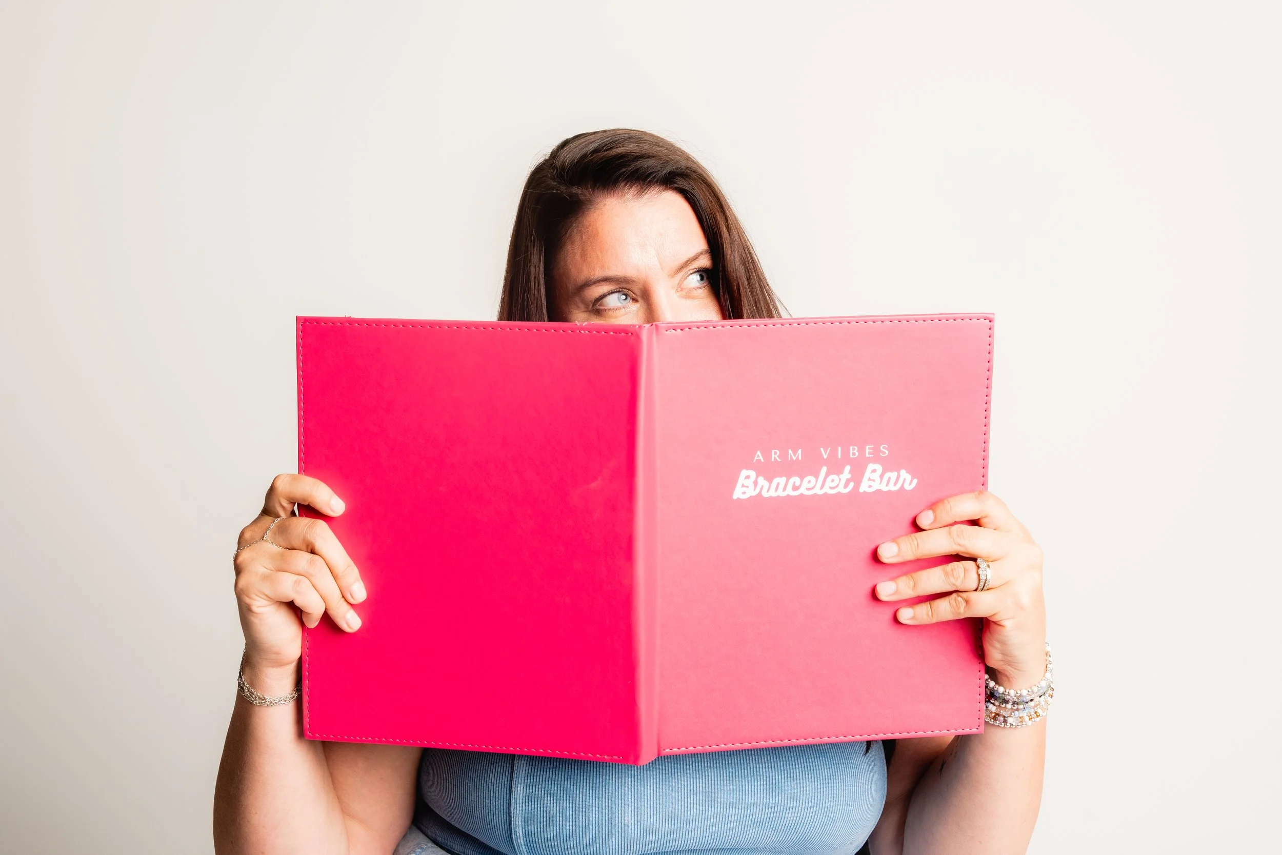 Woman holding a pink book labeled 'Arm Vibes Bracelet Bar' partially covering her face, against a plain white background.