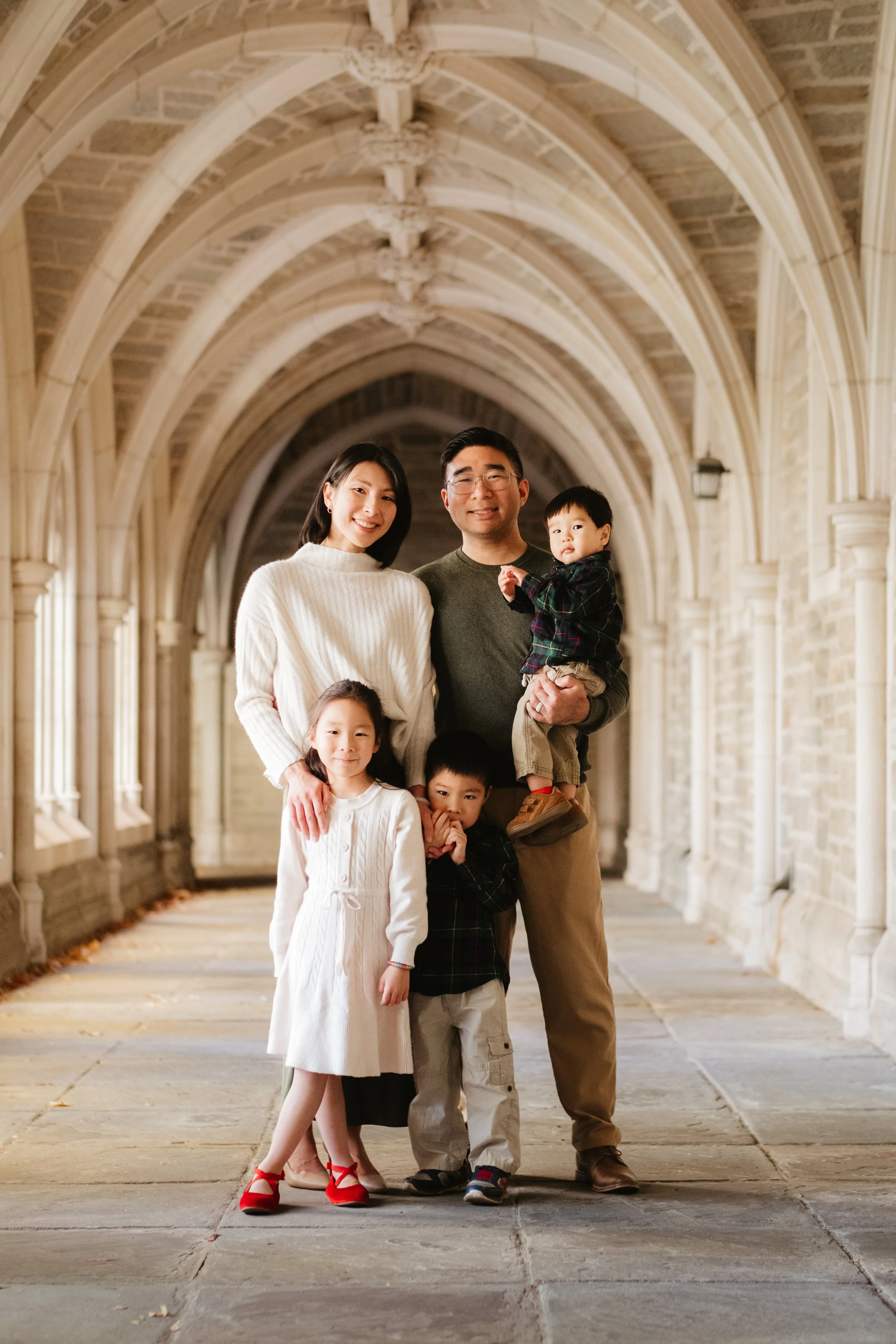 A family of five standing in a stone archway hallway, smiling at the camera. The group includes a woman, a man holding a young boy, a girl, and a boy, dressed in casual clothing.