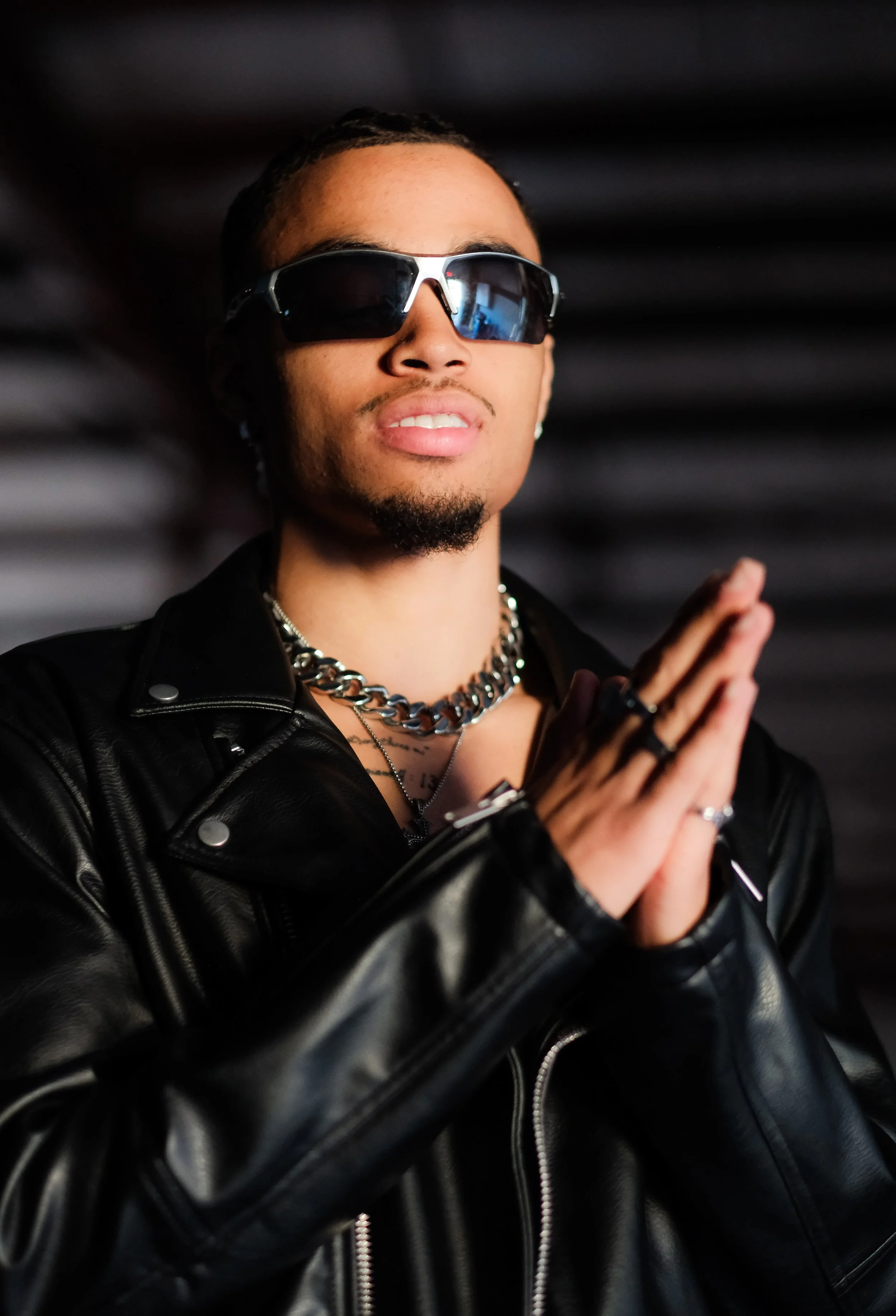 Person wearing sunglasses, chains, a leather jacket, and rings, with hands in a prayer position.
