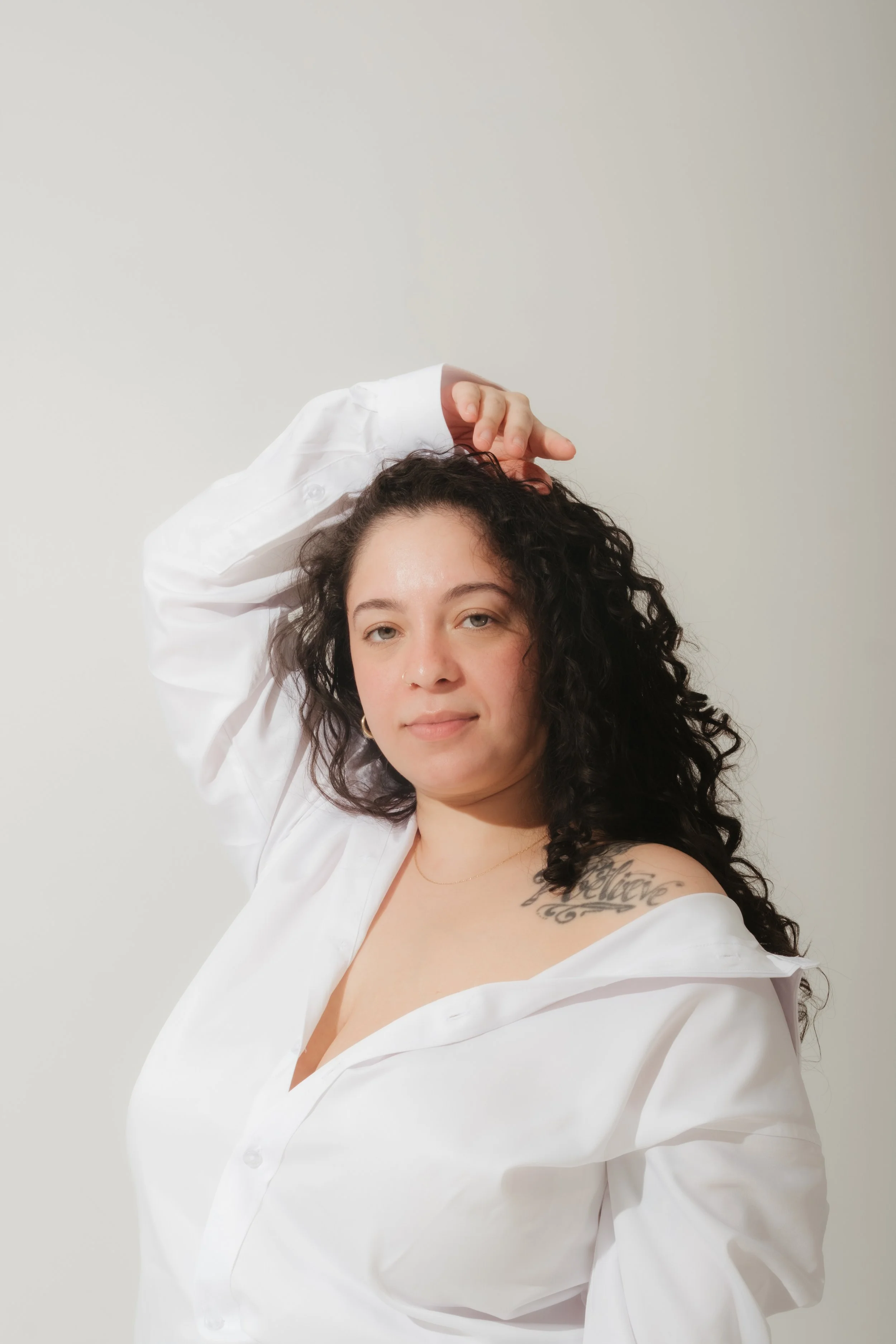 A woman with curly dark hair and a tattoo on her left shoulder poses against a plain white wall, wearing a partially unbuttoned white shirt.