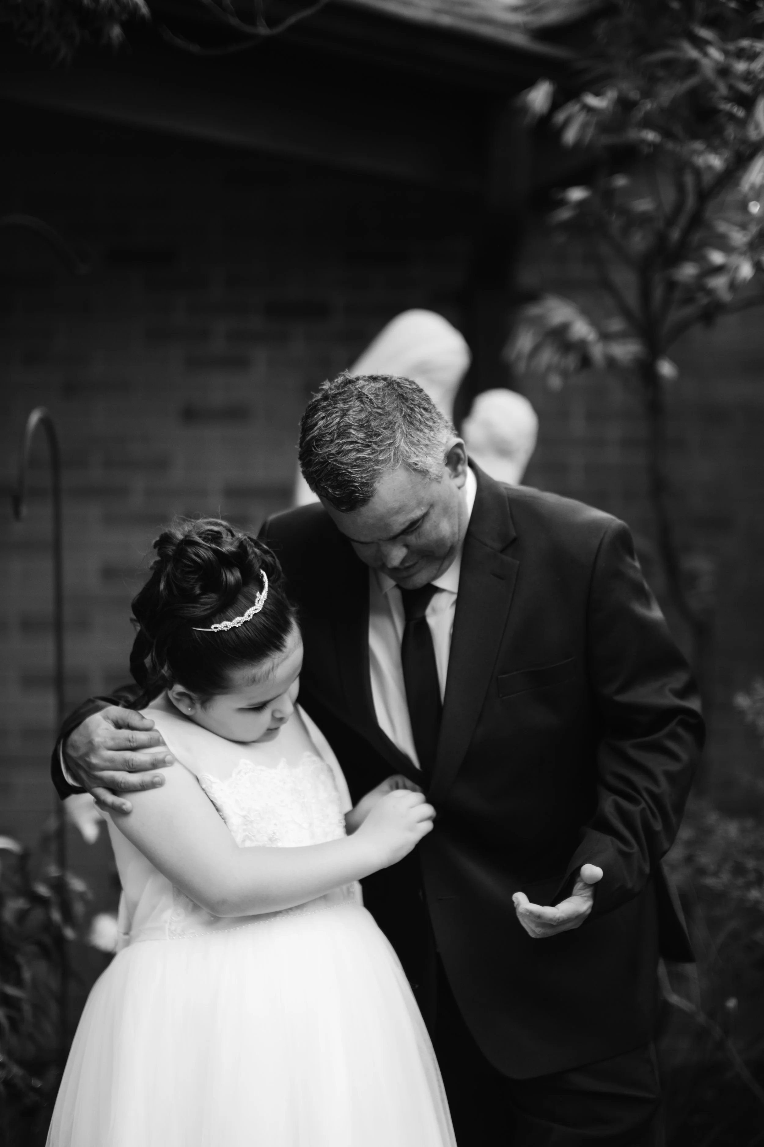 A young girl in a white dress and a headband embraces a man in a suit, both with their heads bowed.