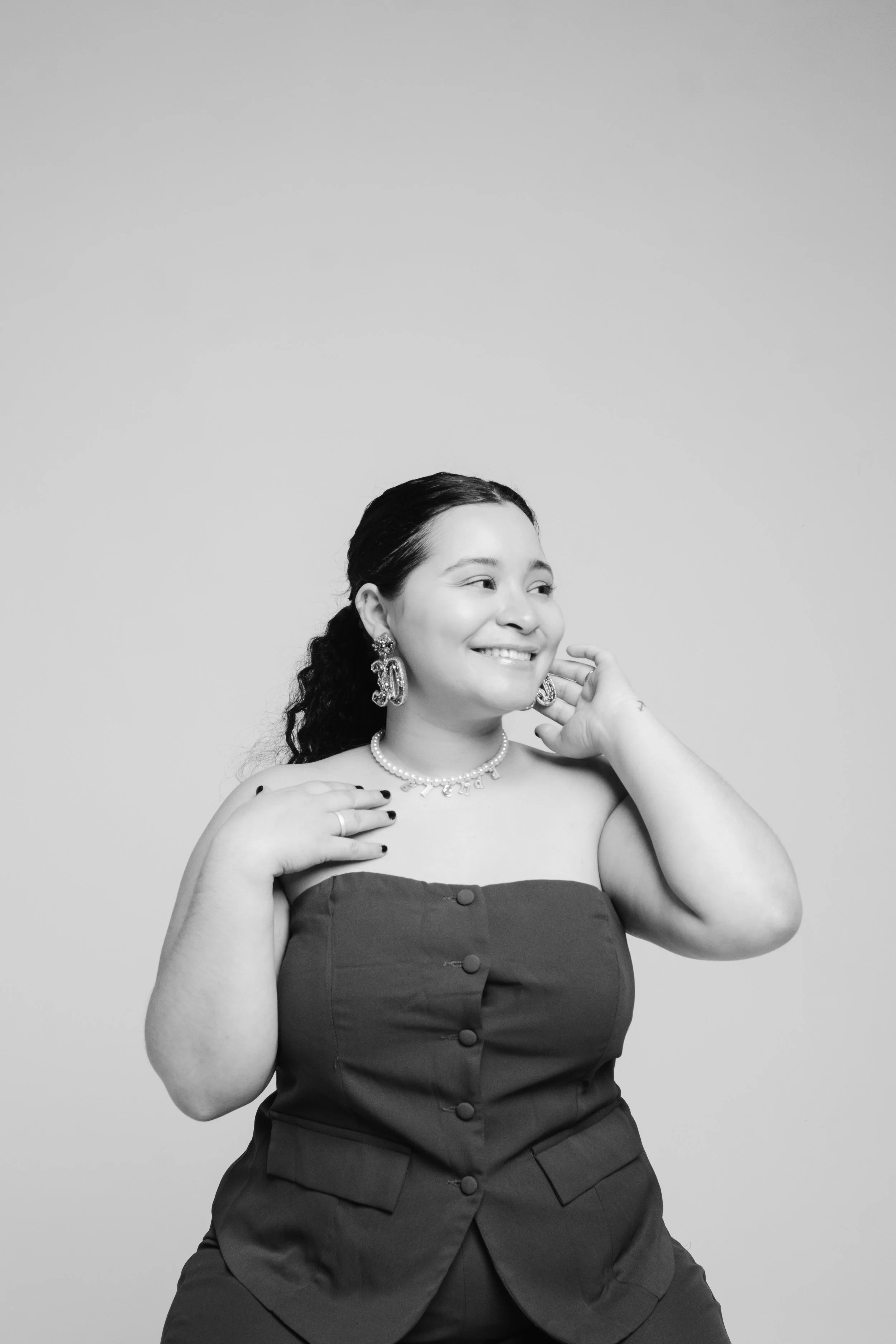 A woman smiling while touching her necklace and earrings, wearing a strapless top, with curly hair and jewelry, against a plain background.