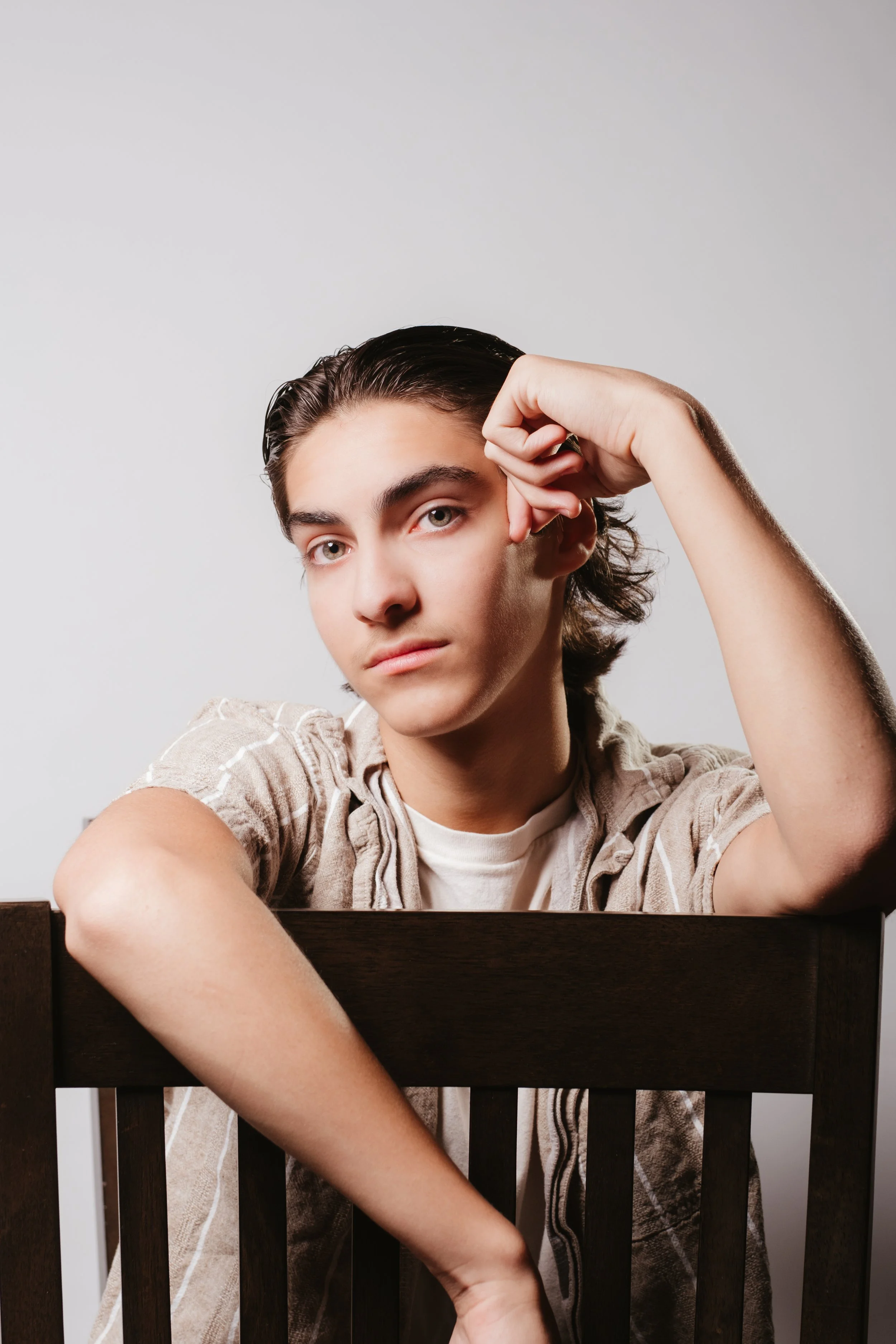 A young man with dark, slicked-back hair and light skin leaning on the back of a wooden chair, resting his cheek on his hand, with a neutral expression, against a plain gray background.