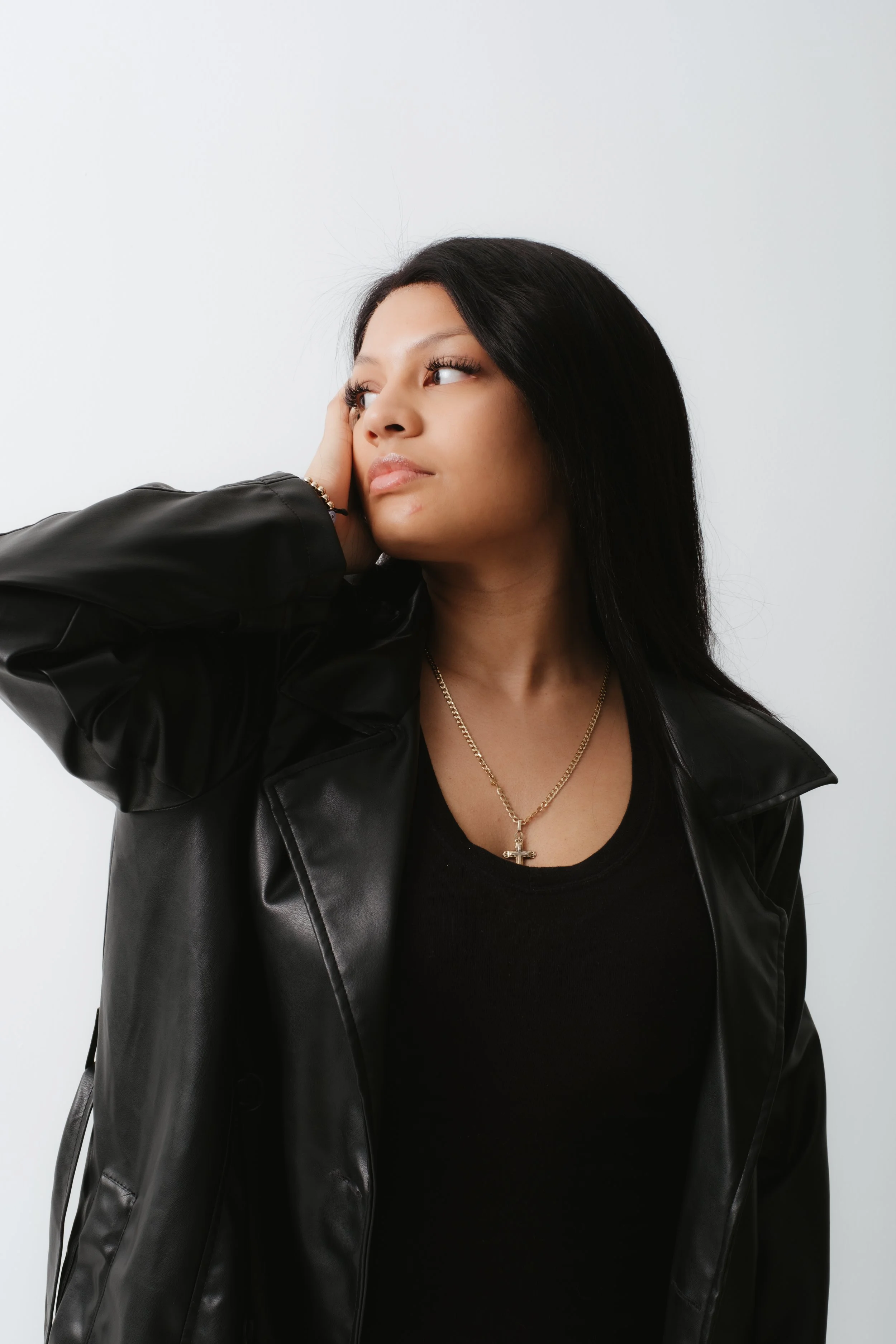A young woman with long black hair, wearing a black leather jacket and a gold cross necklace, gazes to her left with her hand resting on the side of her face, against a plain white background.