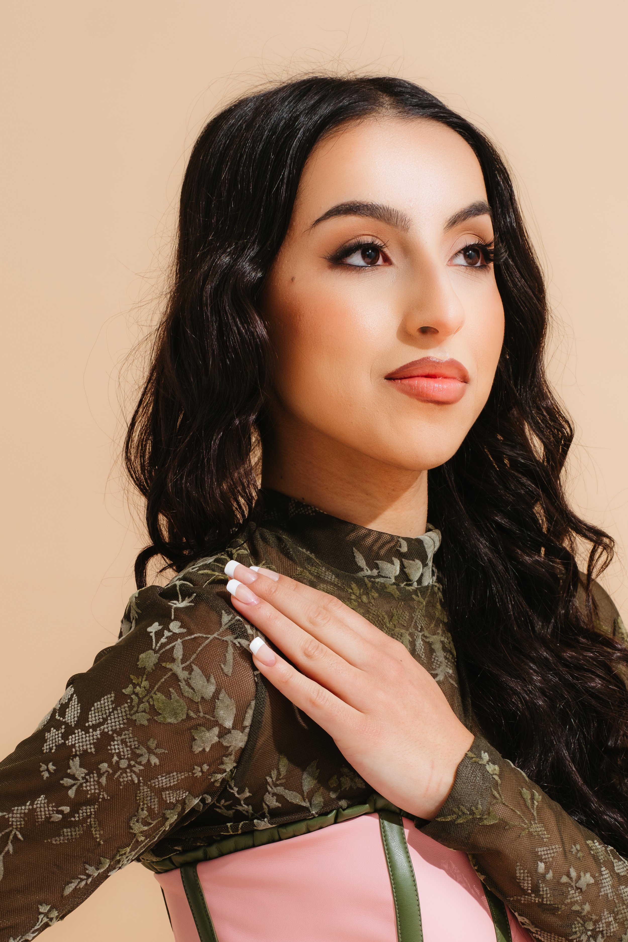 A woman with long, wavy dark hair and makeup, wearing a sheer floral top with green and pink accents, is posing with her hand on her shoulder against a beige background.