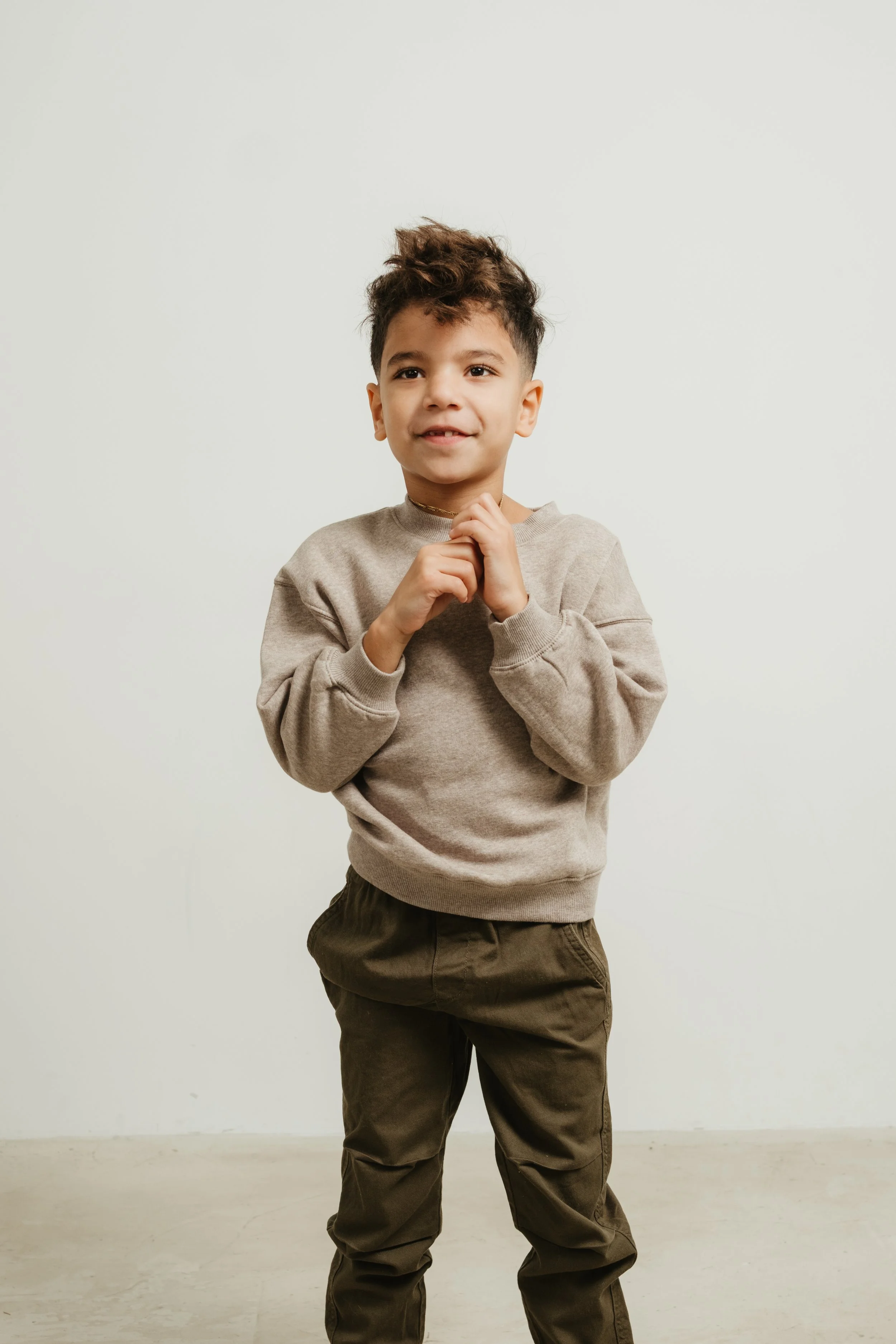 A young boy with curly brown hair, wearing a beige sweatshirt and brown pants, standing against a plain white background with his hands clasped near his chin.