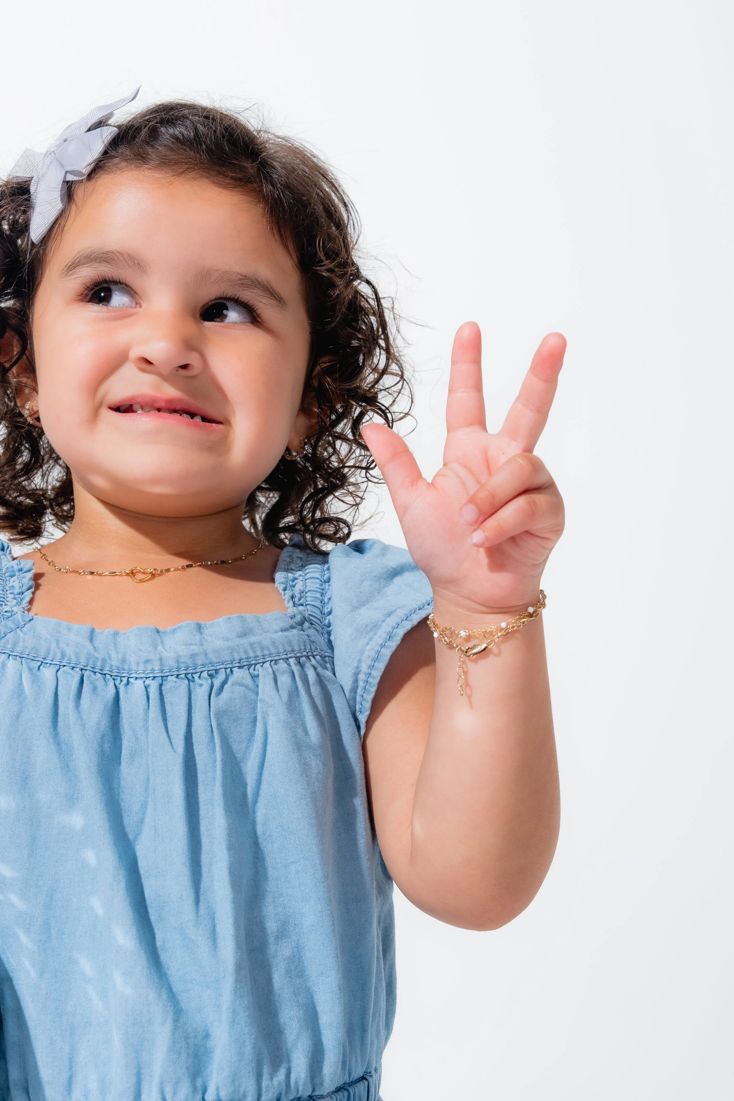 A young girl with curly dark hair, wearing a blue dress and jewelry, making a peace sign with her right hand, looking to her left with a slight smile.