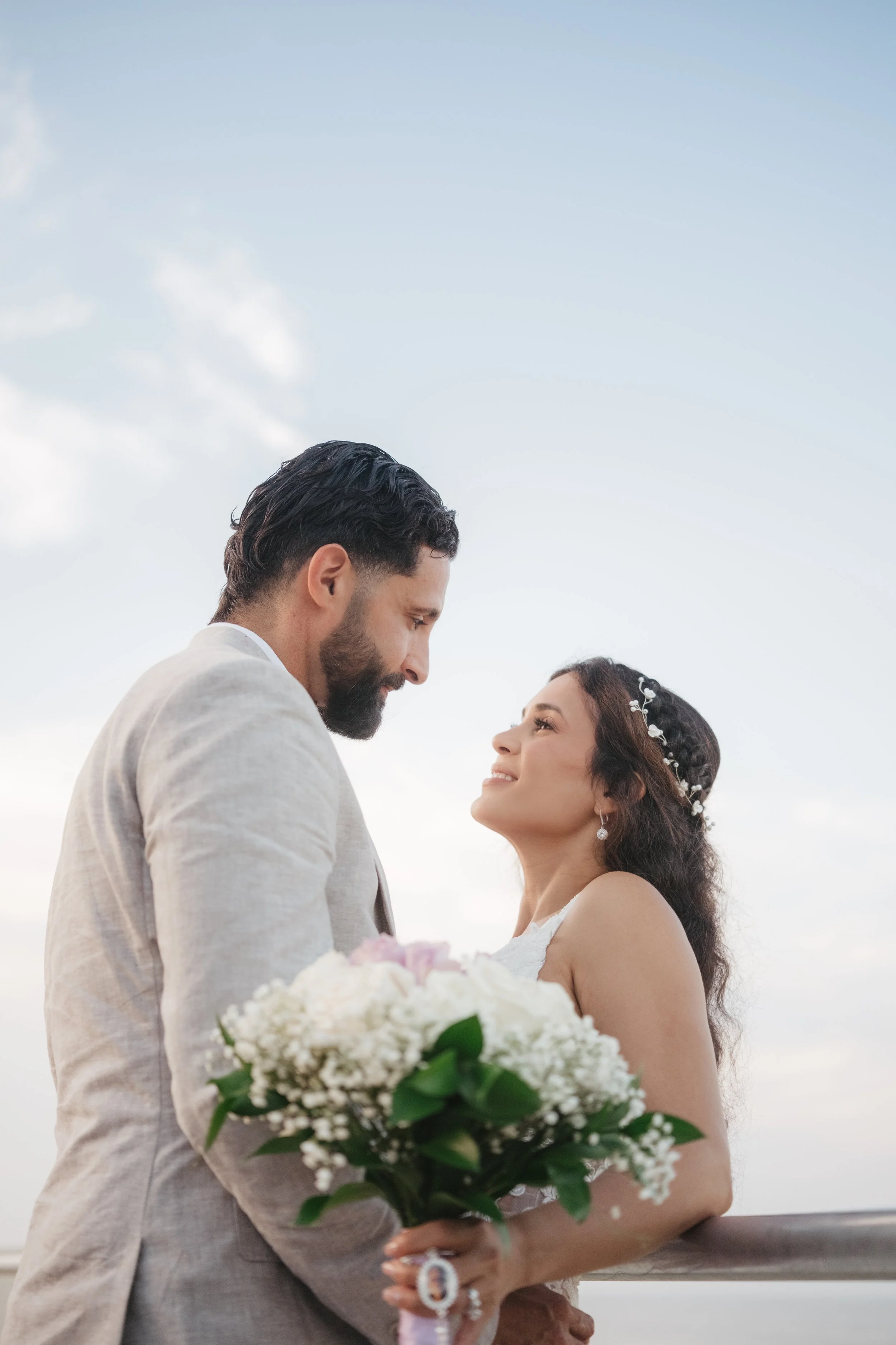 A groom and bride gazing into each other's eyes outdoors with a cloudy sky background. The bride is holding a bouquet of white and pink flowers and wearing a headpiece and wedding dress, the groom is dressed in a light-colored suit.