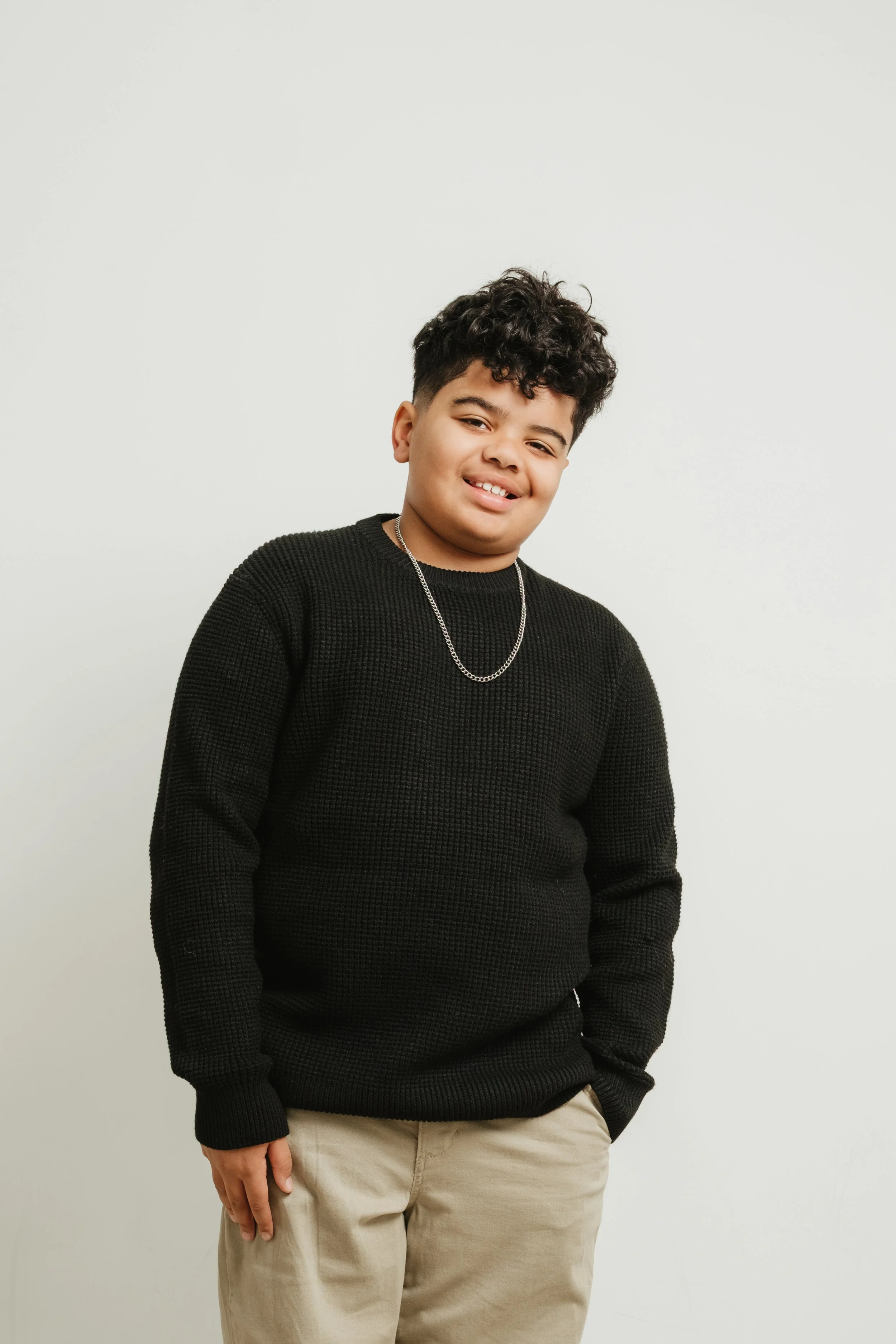 A young person with dark curly hair wearing a black sweater and beige pants, standing against a plain white background, smiling slightly.