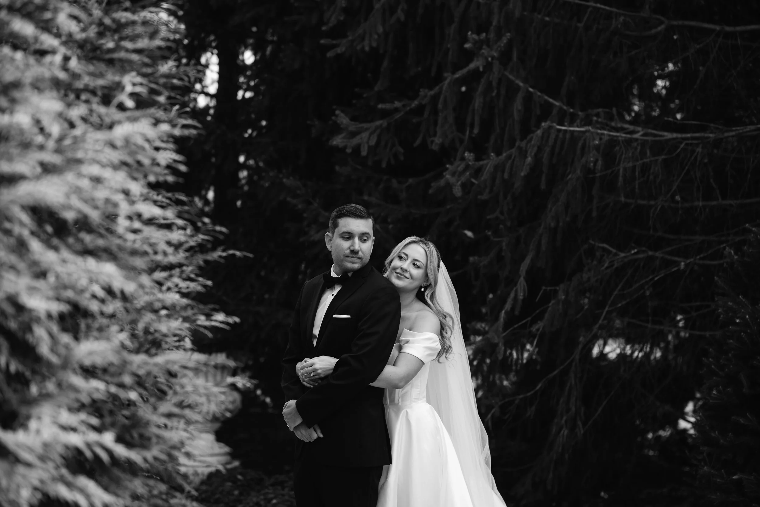 Black and white photo of a bride and groom standing outdoors among trees. The bride is smiling and embracing the groom from behind. The groom is looking to the side.