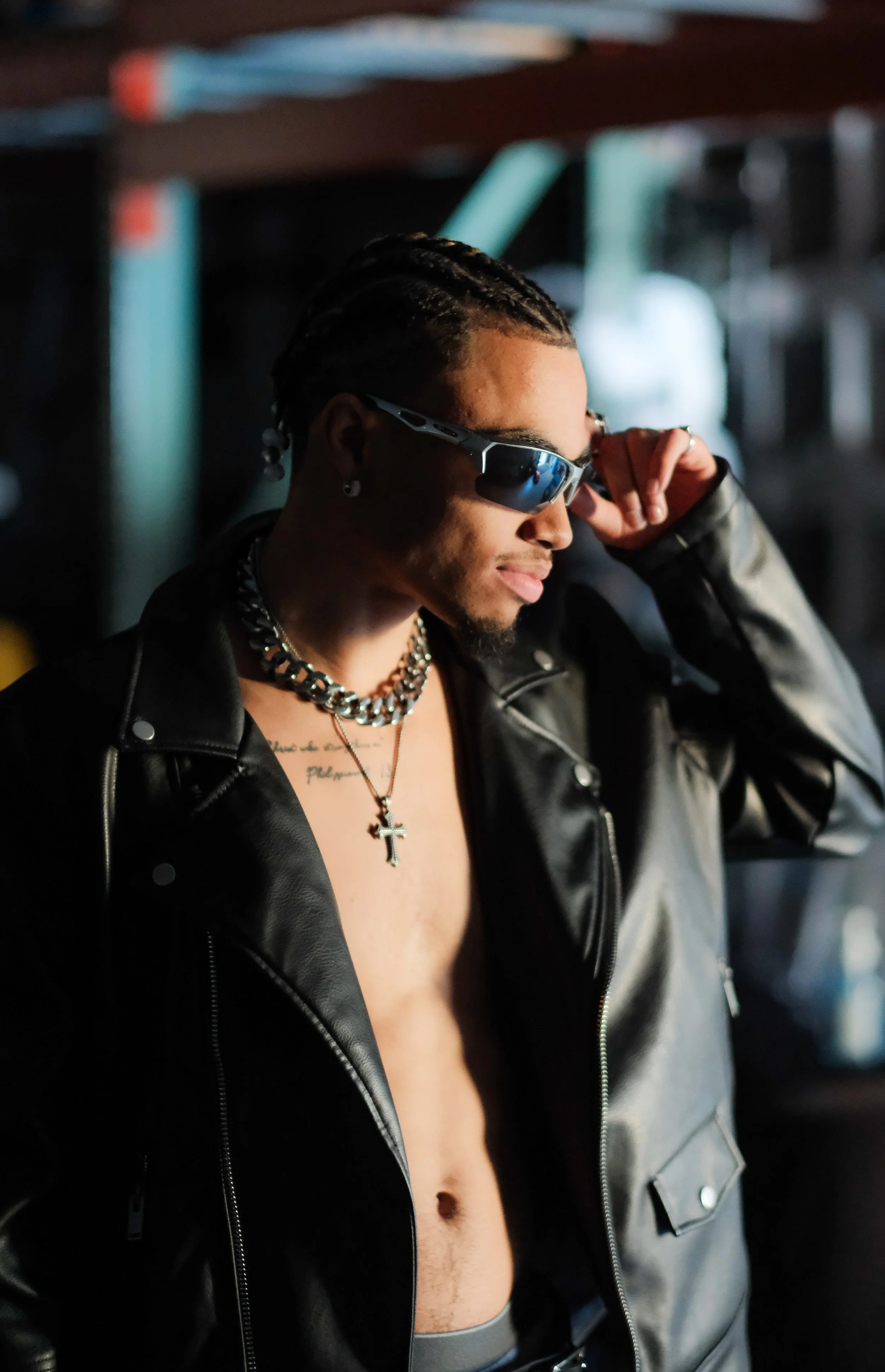 A young man with braided hair, wearing sunglasses, a leather jacket, layered silver necklaces including a cross pendant, and earrings, poses in a dimly lit environment.