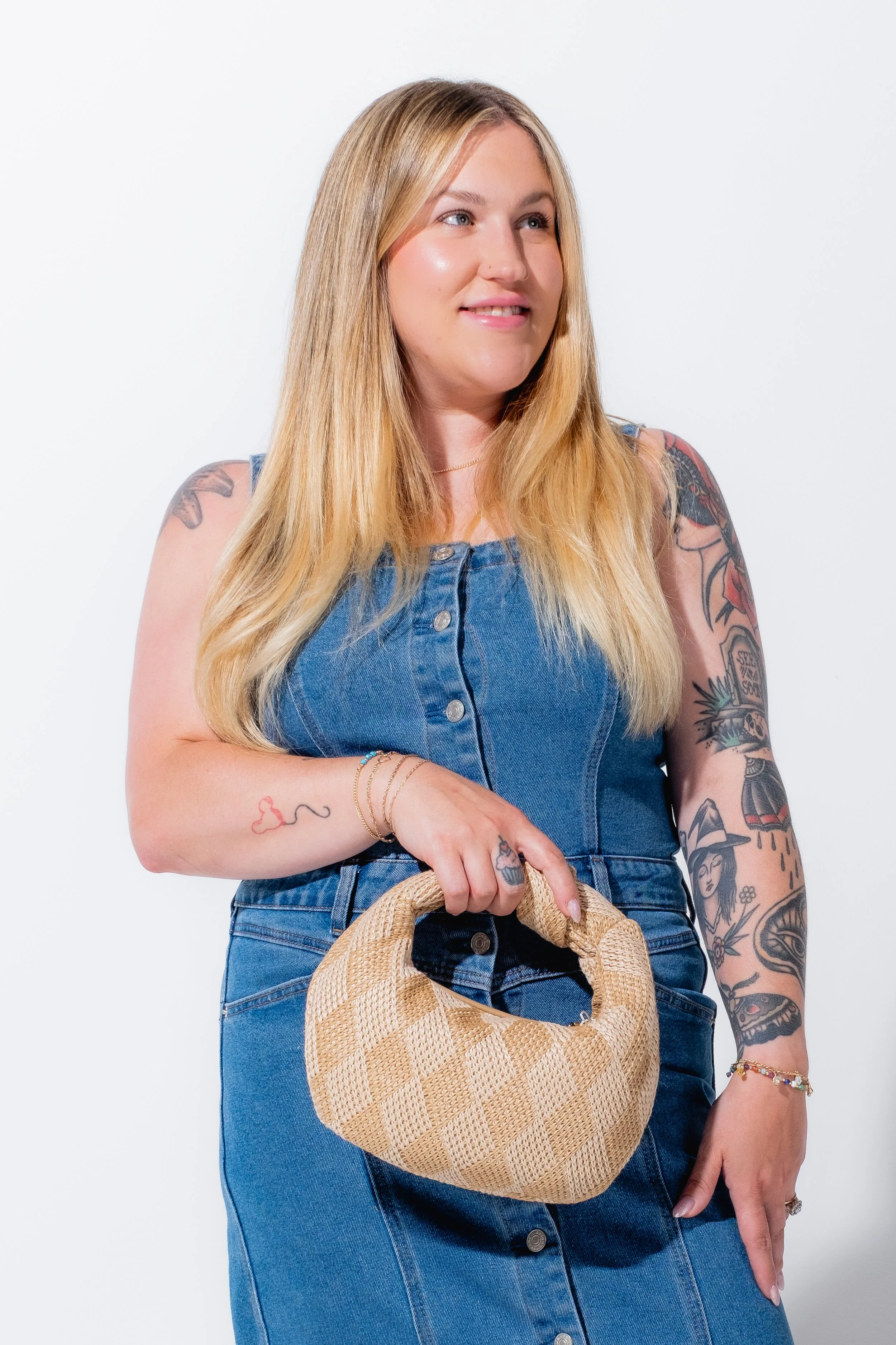 A woman with long blonde hair wearing a sleeveless denim dress, holding a woven beige handbag, standing against a plain white background.