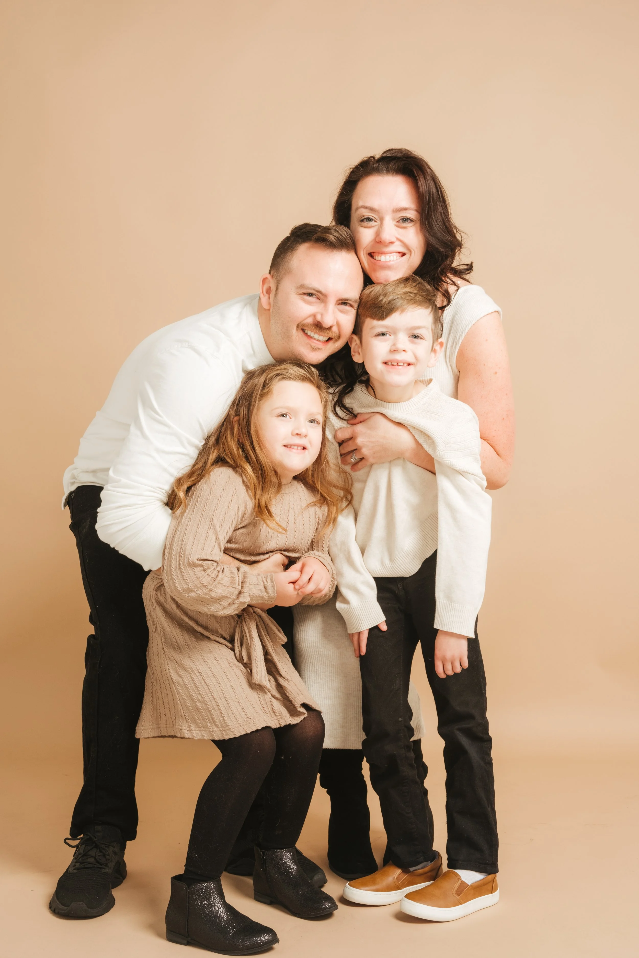 A smiling family of five posing together against a beige background, including a woman, a man, two children, and a young girl, dressed in casual neutral-colored clothing.