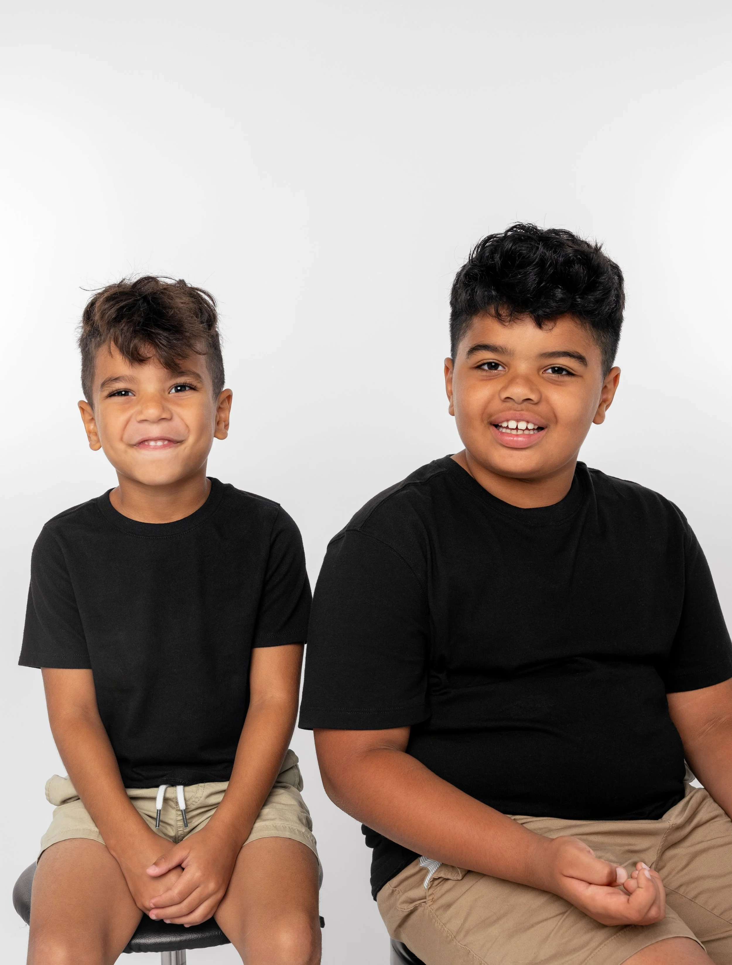 Two boys sitting on chairs against a plain white background, wearing black t-shirts and beige shorts, smiling at the camera.