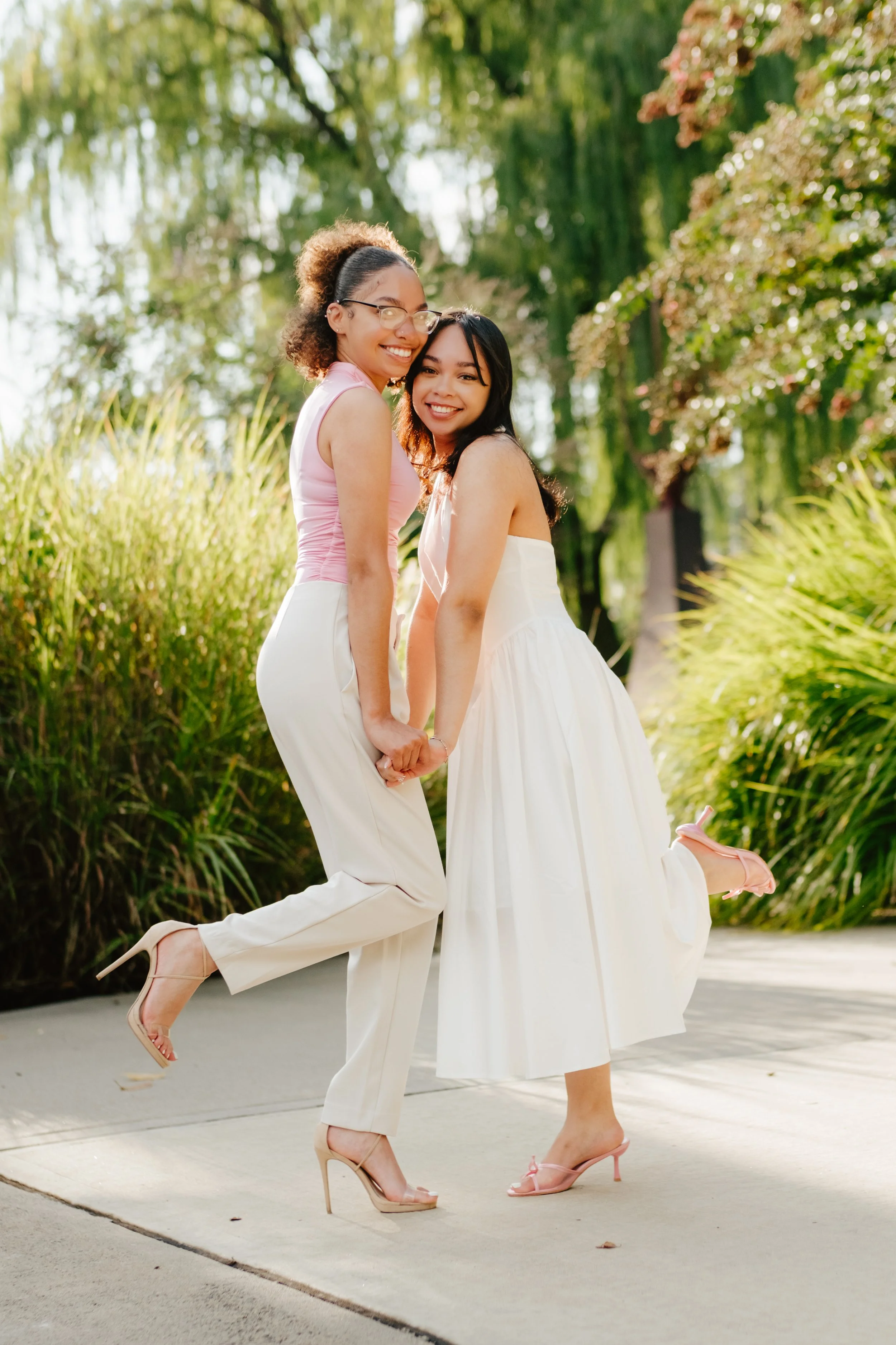 Two women holding hands, lifting one leg in a park with trees and tall grass, both wearing white dresses and pink heels, smiling at the camera.