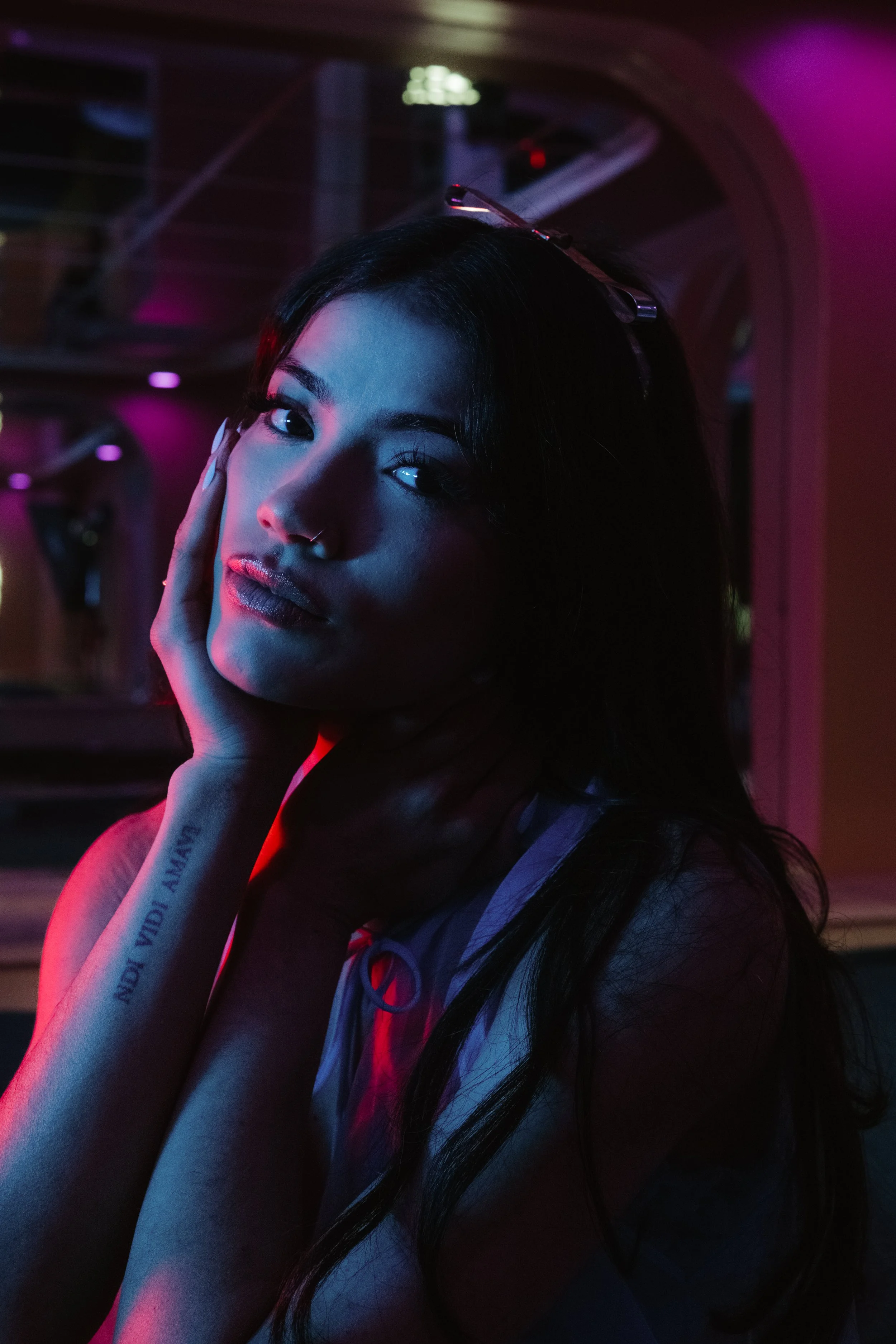 A woman with dark hair and a nose ring resting her face in her hand, posed in a dimly lit room with colorful neon lighting.