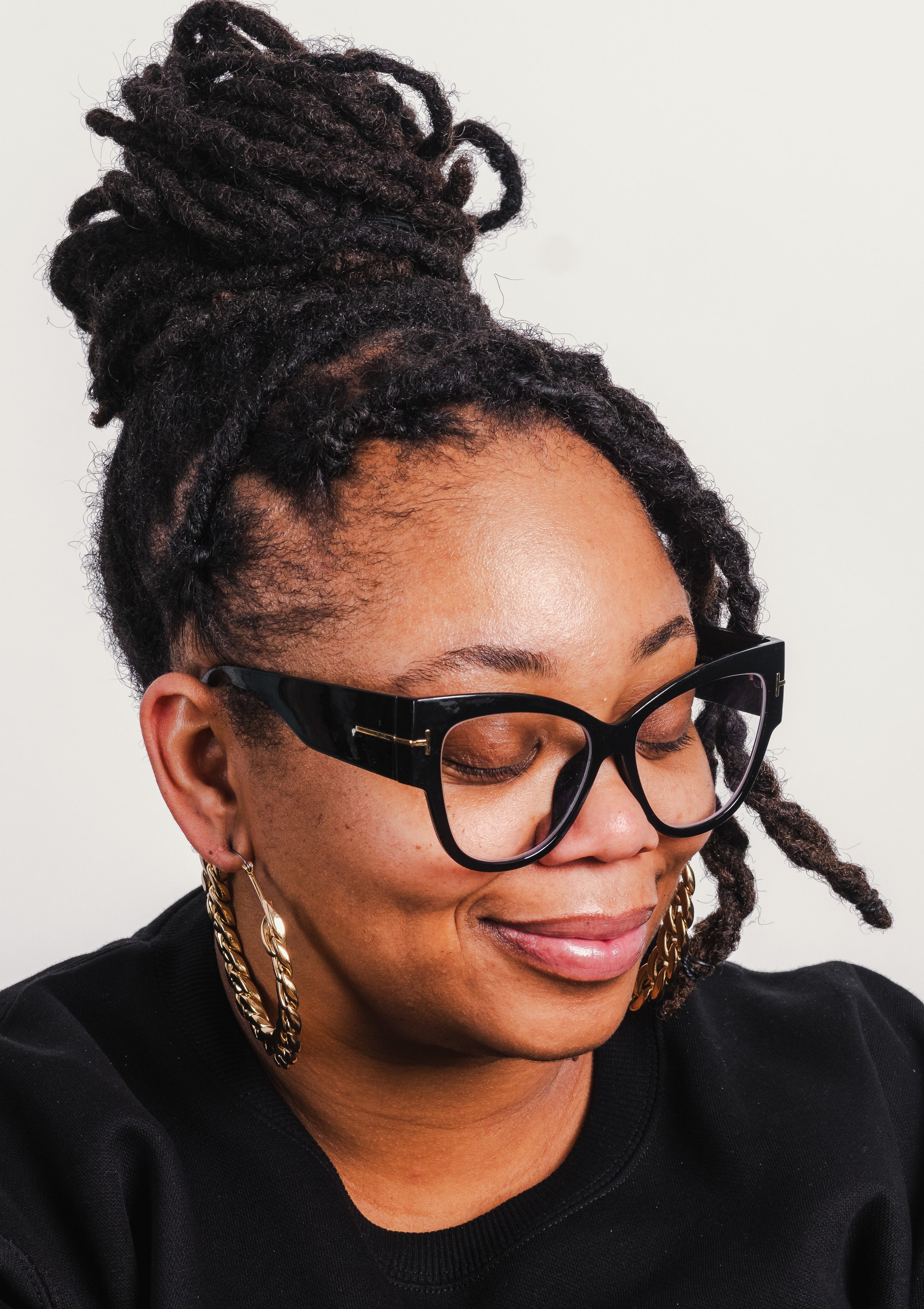A woman with styled dreadlocks in a high bun, wearing black glasses with gold accents, hoop earrings, and a black top, with her eyes closed and a gentle smile.