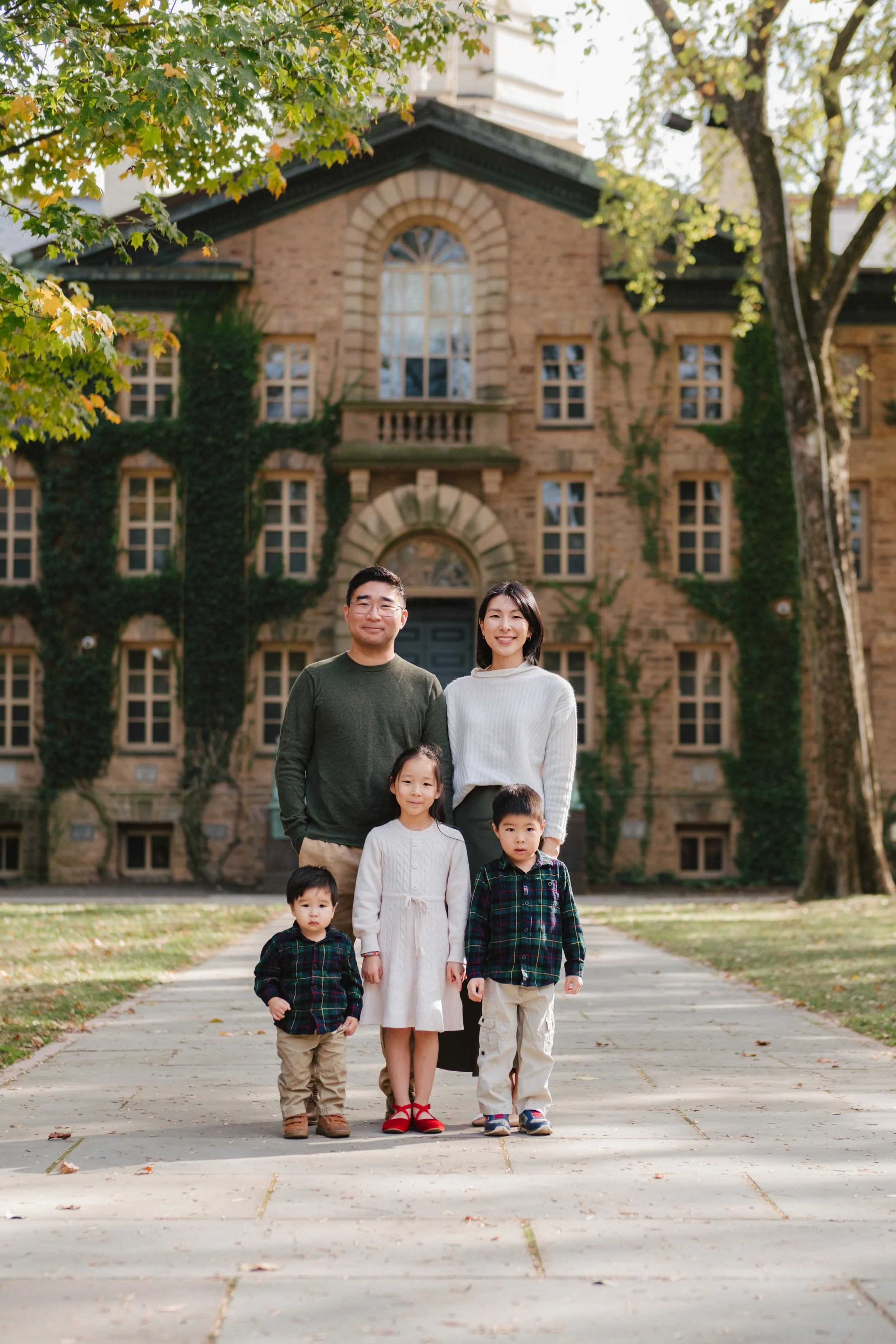 A family of six posing on a sidewalk in front of a large brick house with ivy-covered walls and trees with autumn leaves.