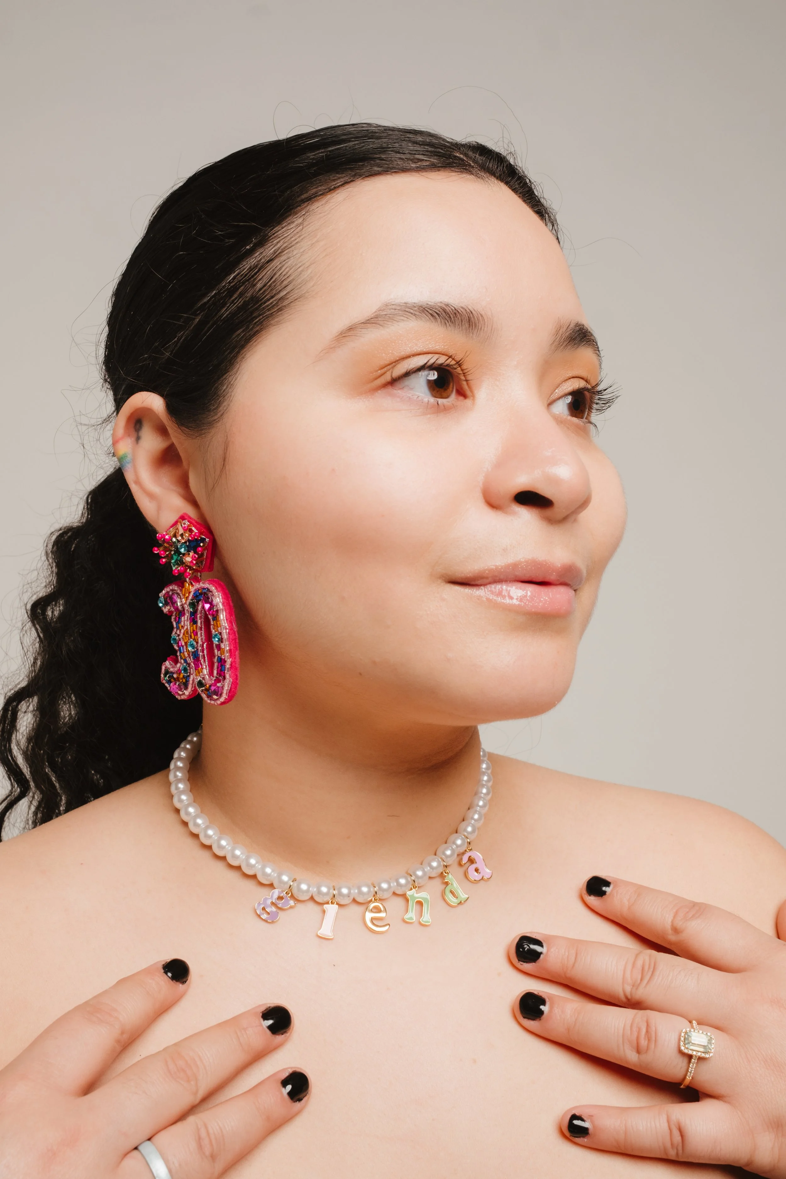 Close-up of a woman with dark hair pulled back, wearing colorful earrings, a pearl necklace with the name 'Fenda' on it, and a ring with a large rectangular stone. She has black nail polish and is touching her collarbone with both hands, looking slig