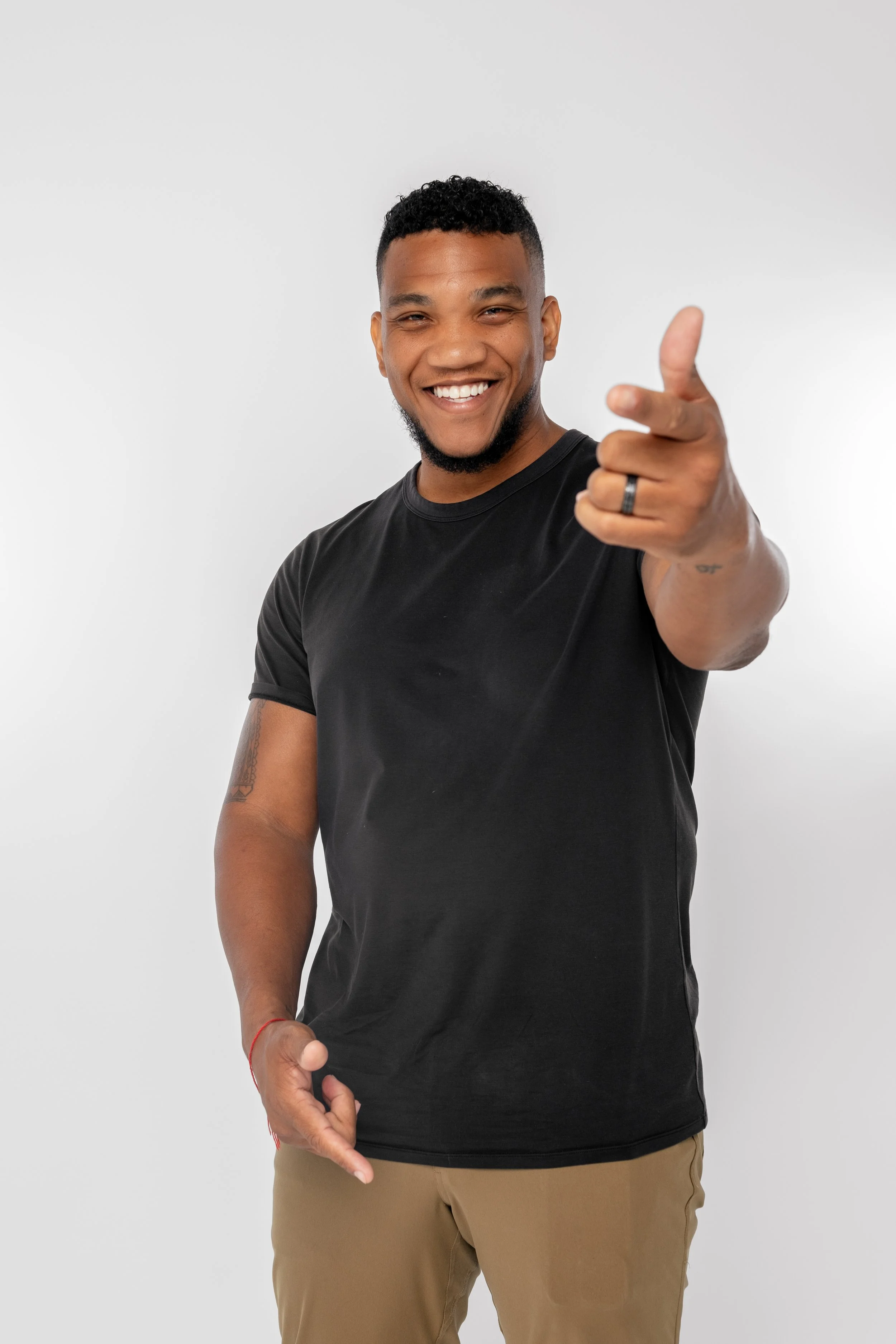 A smiling young man with a beard and short curly hair, wearing a black t-shirt and beige pants, pointing towards the camera against a plain white background.