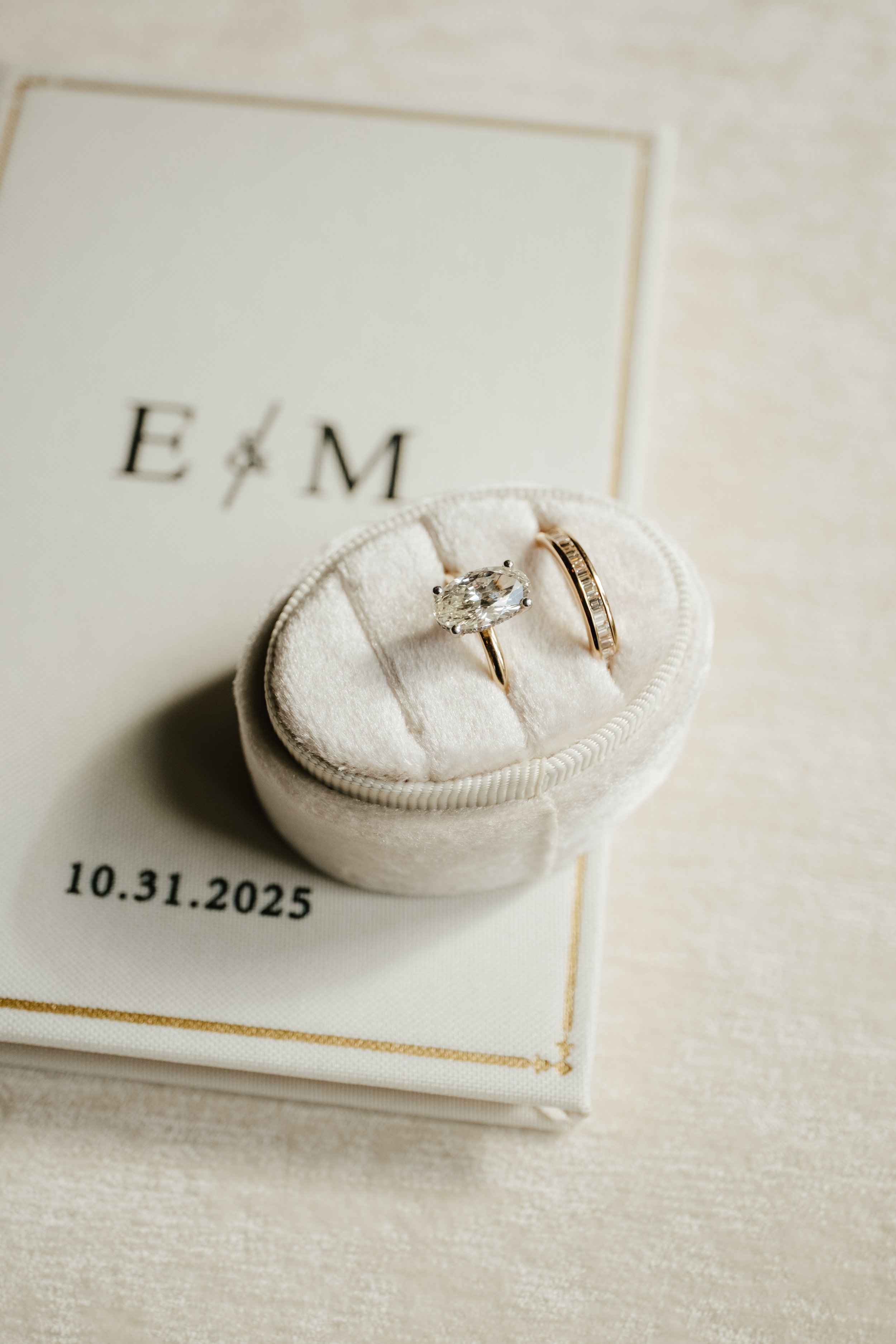 A jewelry box containing two rings, one with a large oval diamond and the other with smaller diamonds, placed on a white cloth with a wedding date of October 31, 2025, in the background.