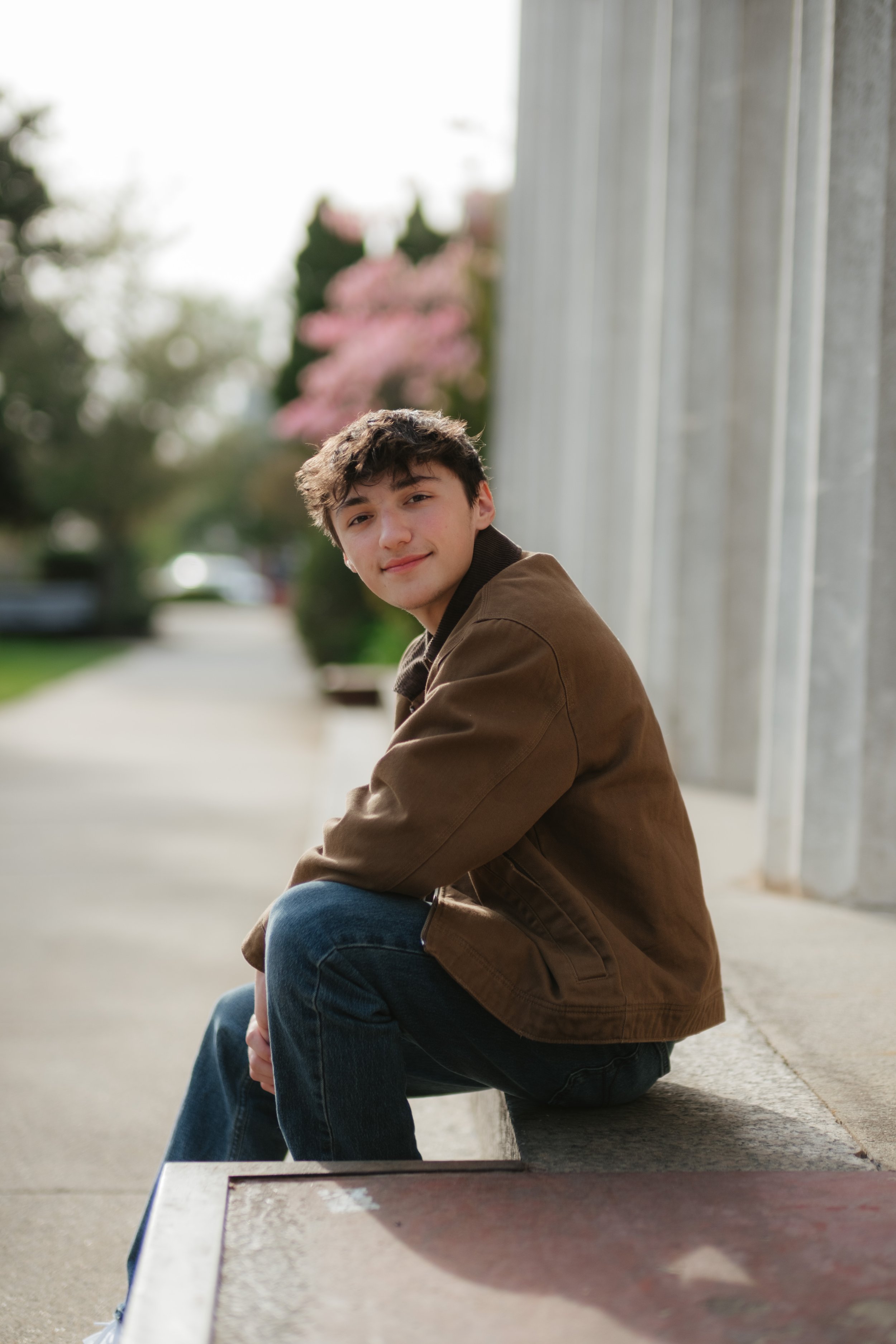 A young man with dark, curly hair, wearing a brown jacket and blue jeans, sitting on a ledge outdoors near a building with columns, with trees and pink blossoms in the background.