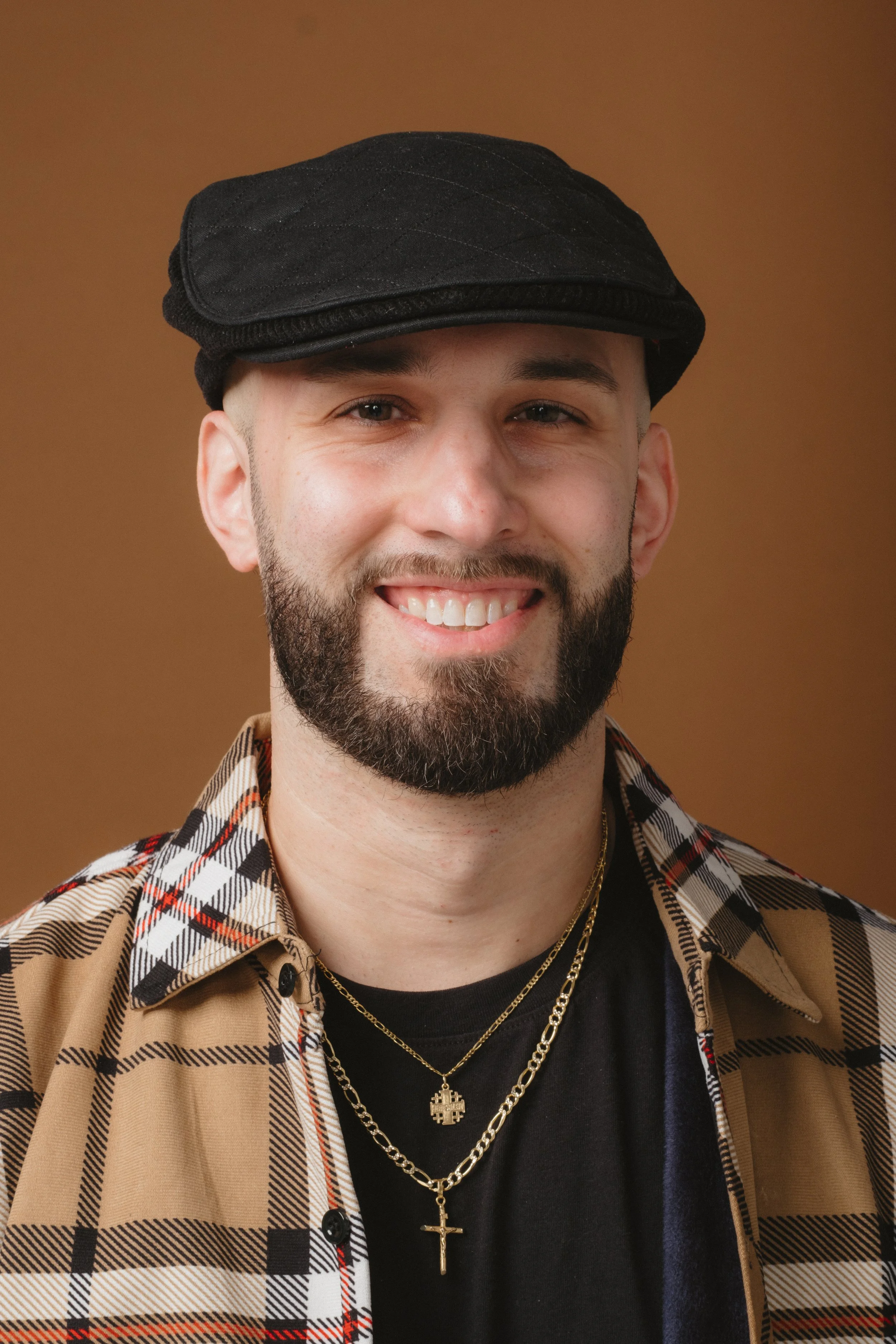 A smiling man with a beard, wearing a black cap, layered gold necklaces, a black T-shirt, and a plaid button-up shirt, against a brown background.