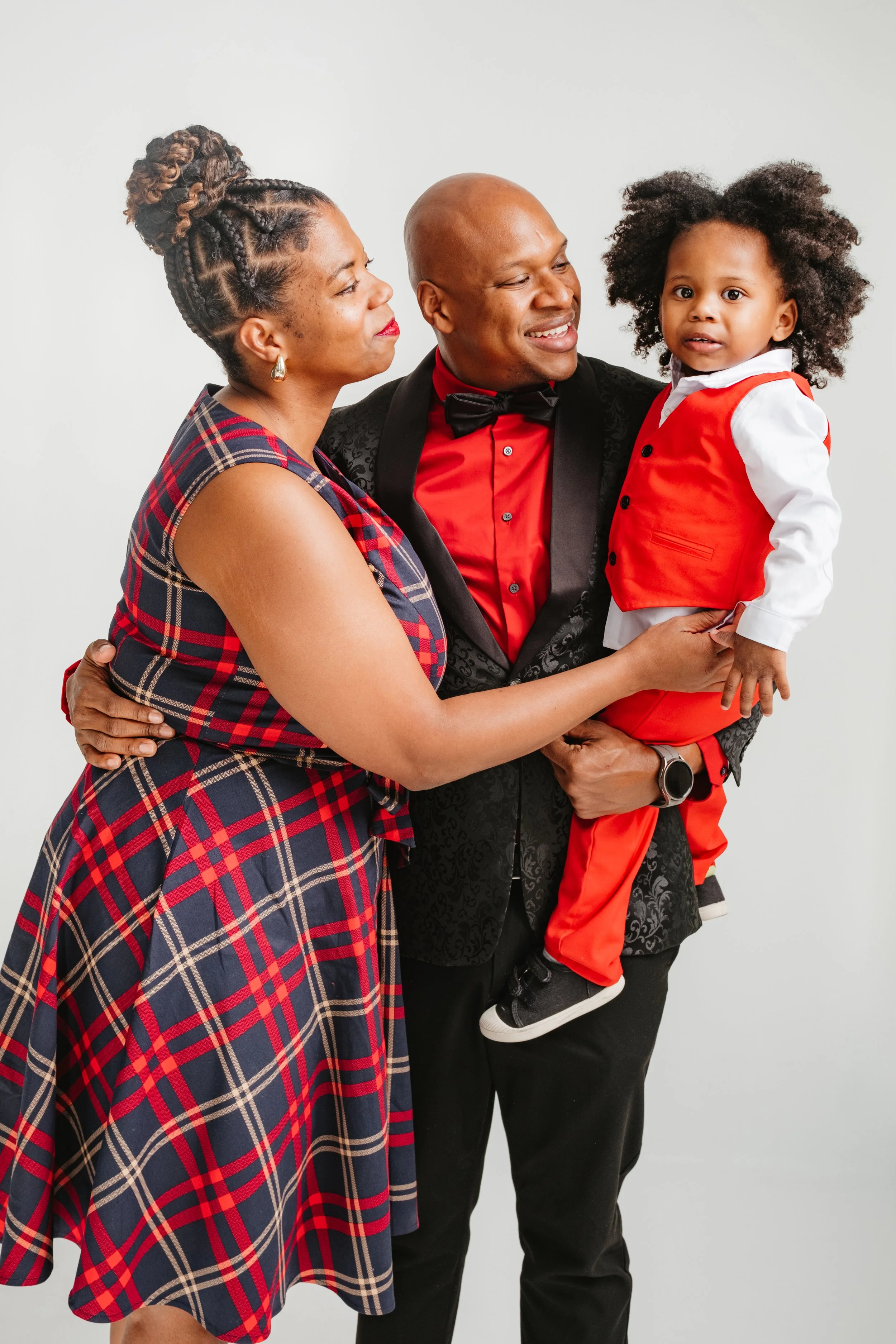 A family of three celebrating. The woman is wearing a plaid dress, the man is dressed in a tuxedo with a red shirt and bow tie, and the young boy is in a red vest and pants with a white shirt. They are smiling and looking at each other, with the woma