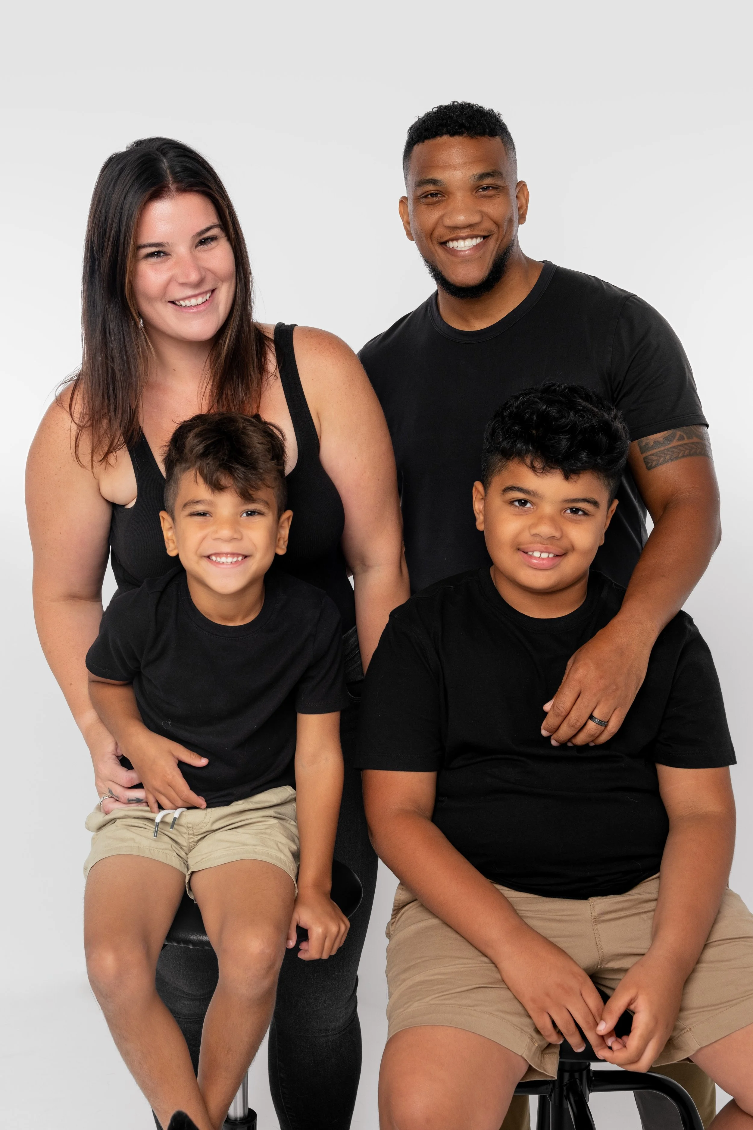 A family of four smiling against a white background. The woman has long dark hair and is wearing a black top. The man has short curly hair, a beard, and is wearing a black T-shirt. They have two boys sitting in front; both are wearing black T-shirts 