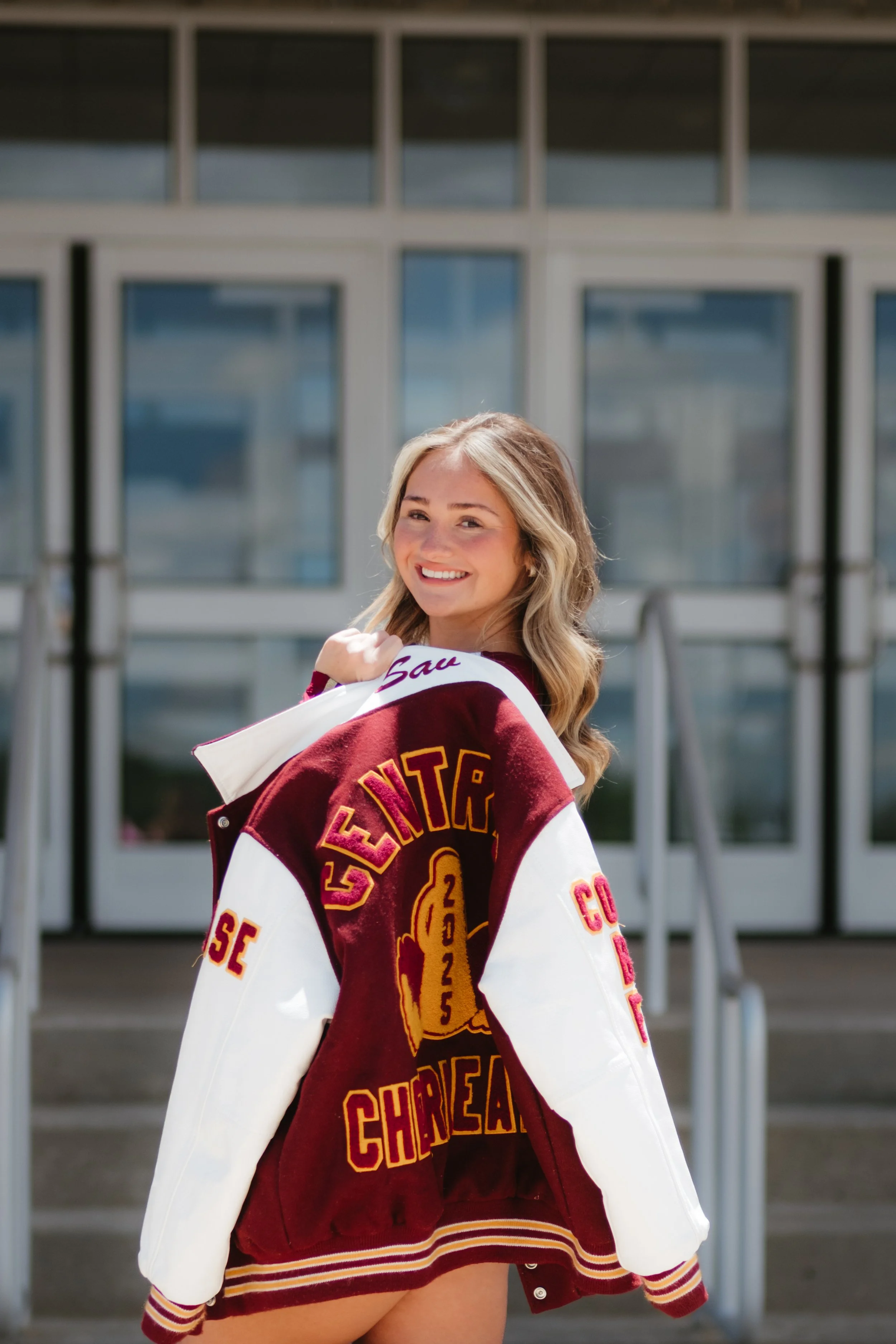 Young woman smiling while wearing a varsity jacket with 'Glenview Chargers' embroidered on the back, standing outdoors in front of a modern building with glass doors.