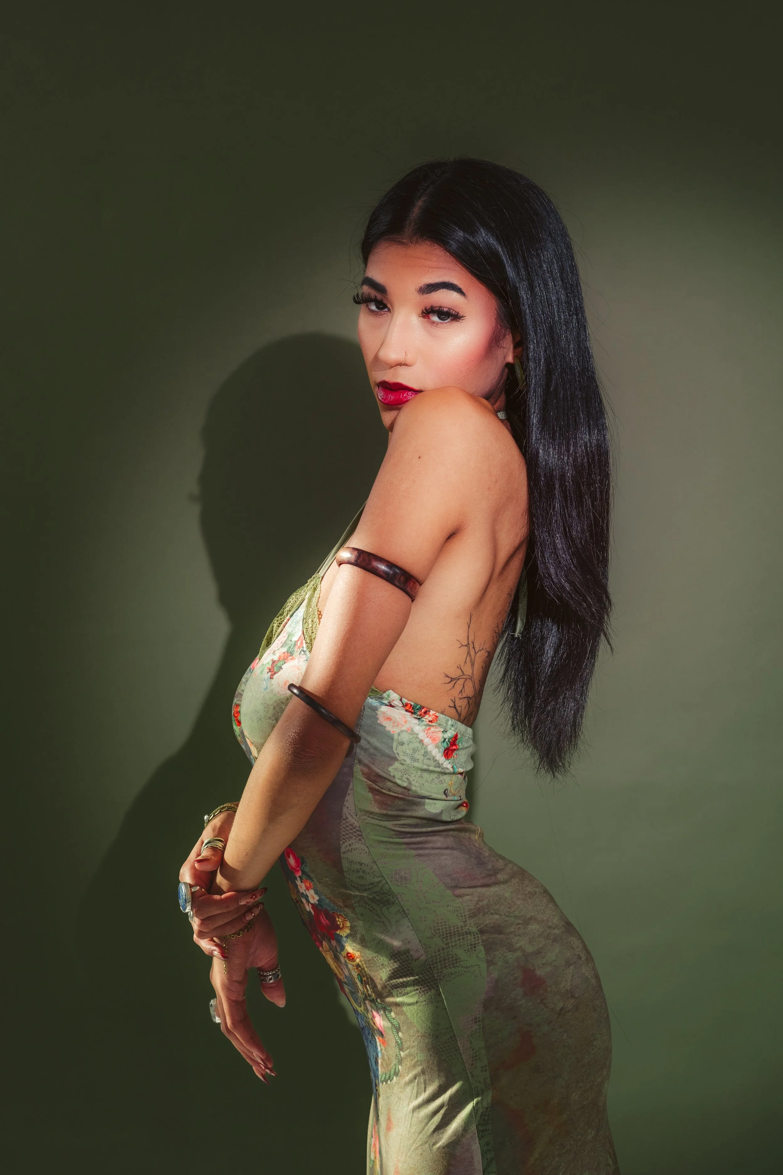 A young woman with long dark hair, wearing a floral dress, standing against a green background. She has a confident expression and her arms are adorned with jewelry.