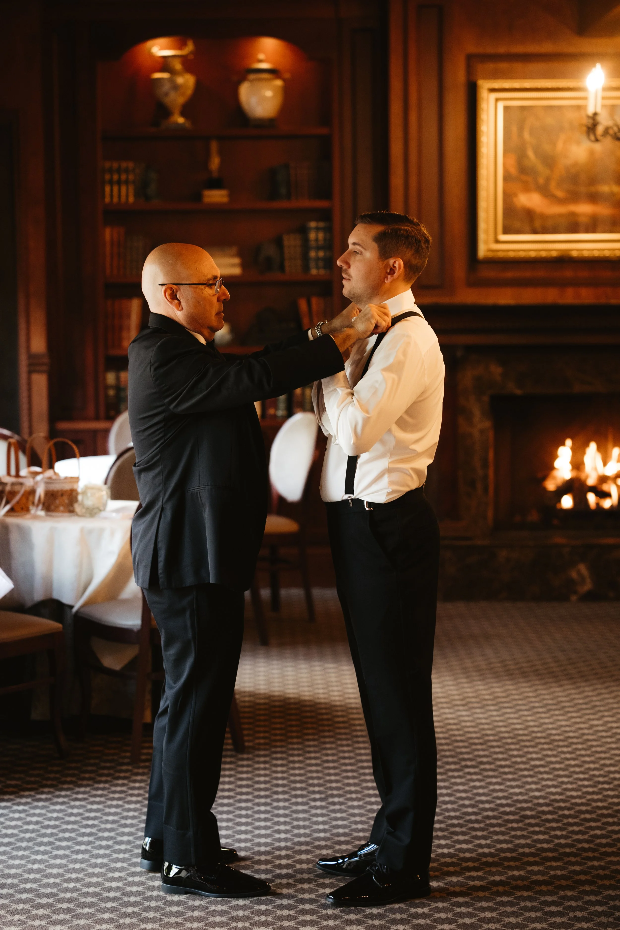An older man adjusting a younger man’s tie in a warmly lit, elegant room with a fireplace and bookshelves.