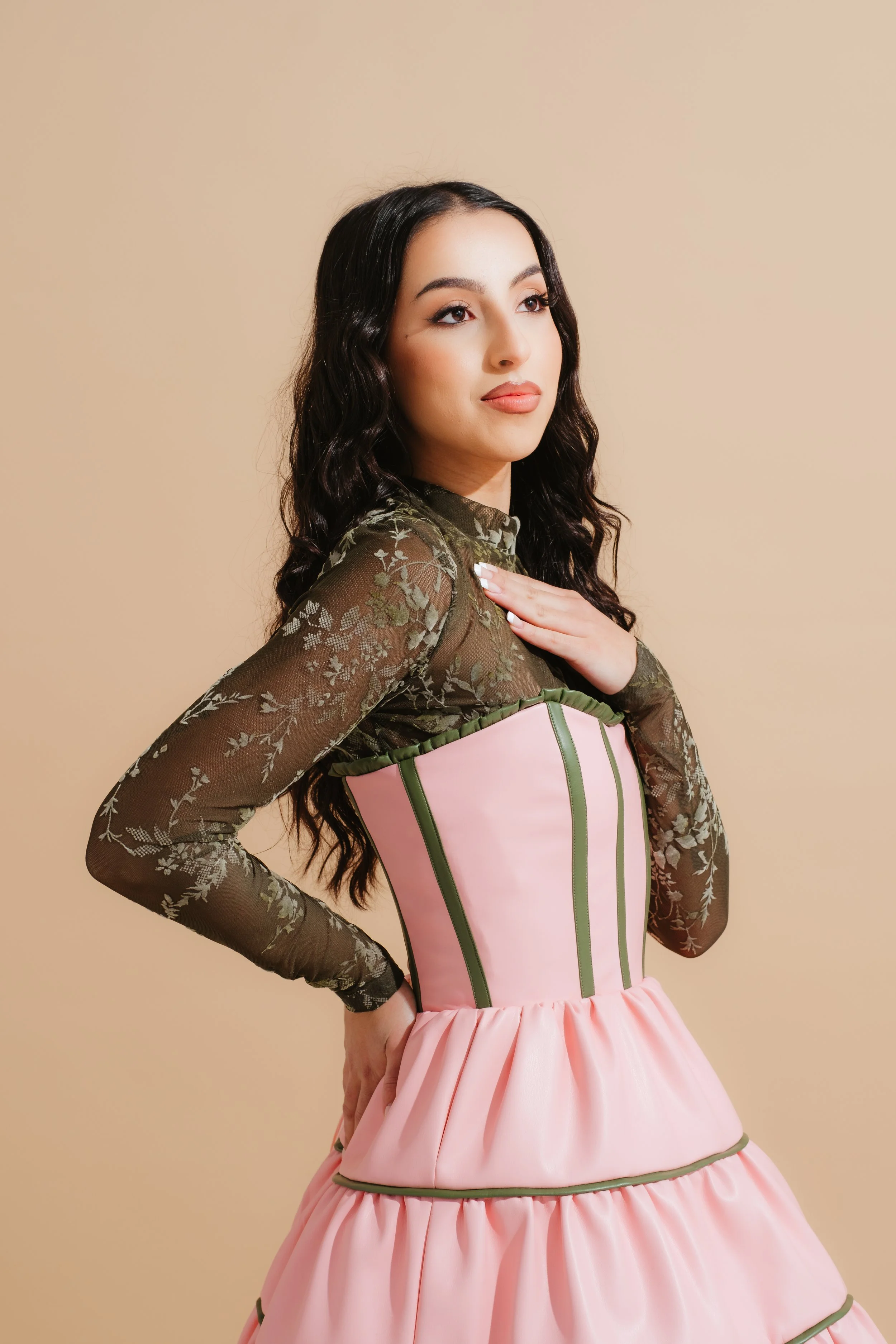 A young woman with long, dark, wavy hair, wearing a sheer, long-sleeved, green lace top and a pink dress with green piping, posing with her hand on her chest against a beige background.