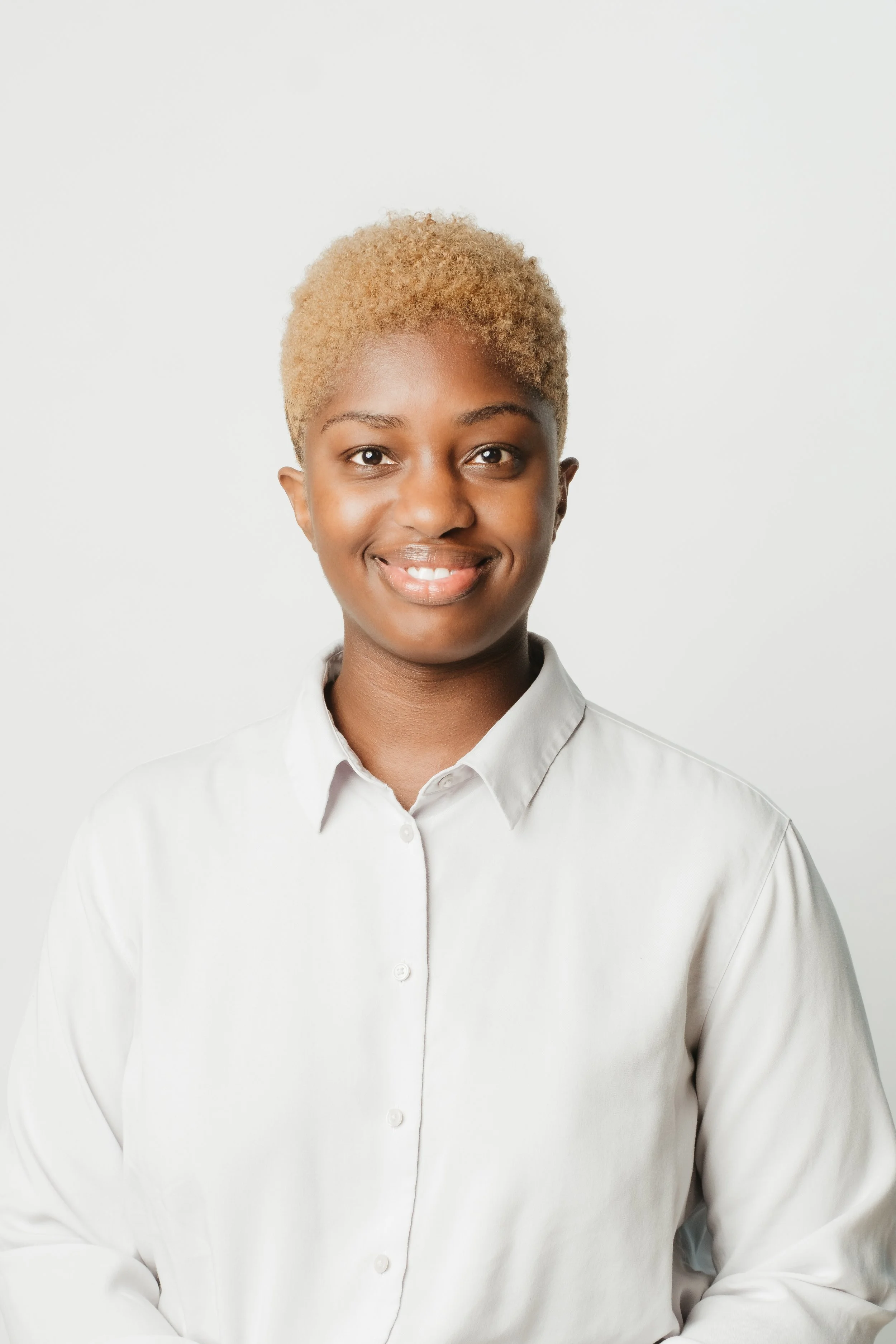 Portrait of a young woman with short blonde hair, smiling, wearing a white collared shirt, against a plain white background.