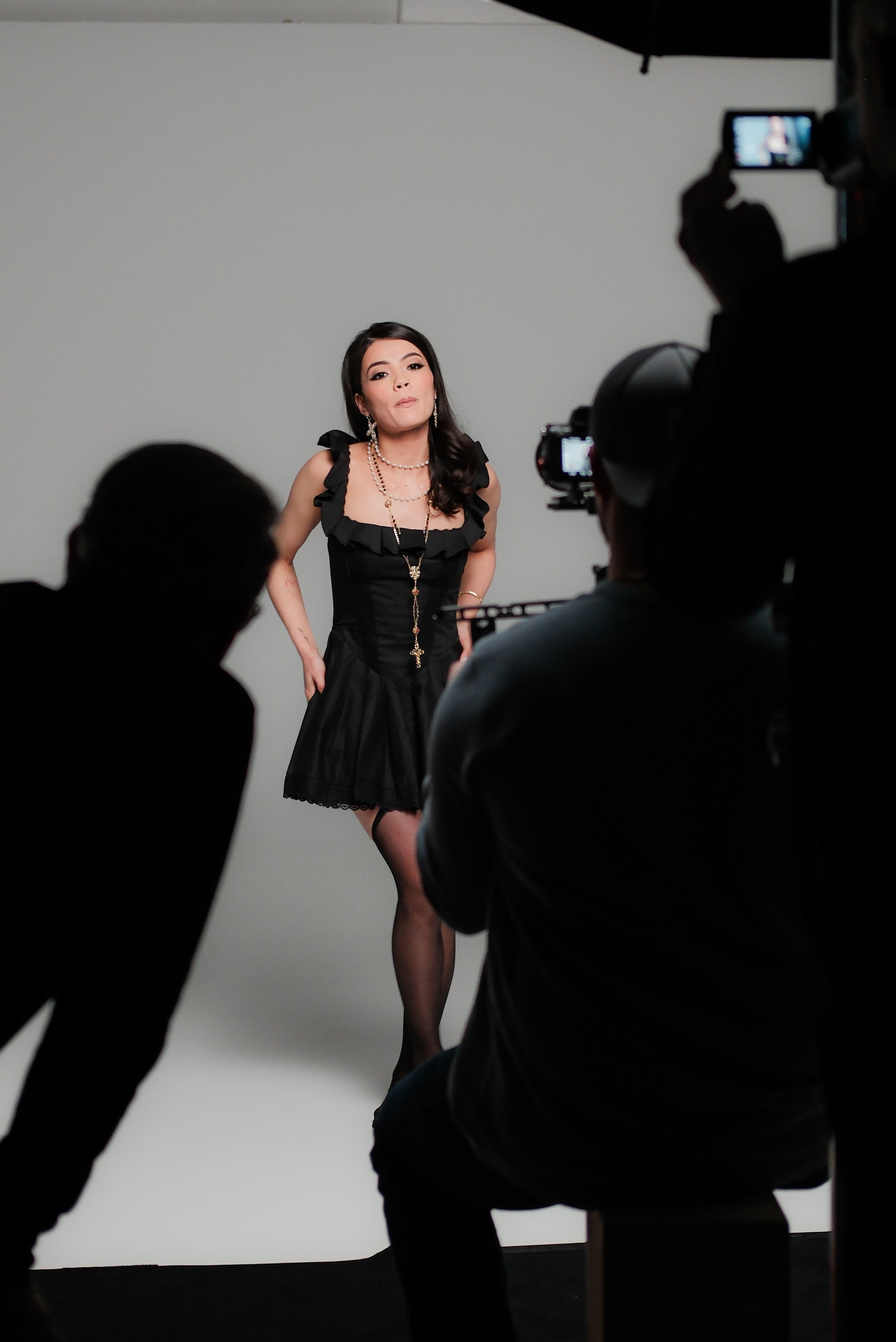 A woman in a black dress and jewelry posing for a photoshoot in a studio with a white backdrop, surrounded by photographers capturing her photos.