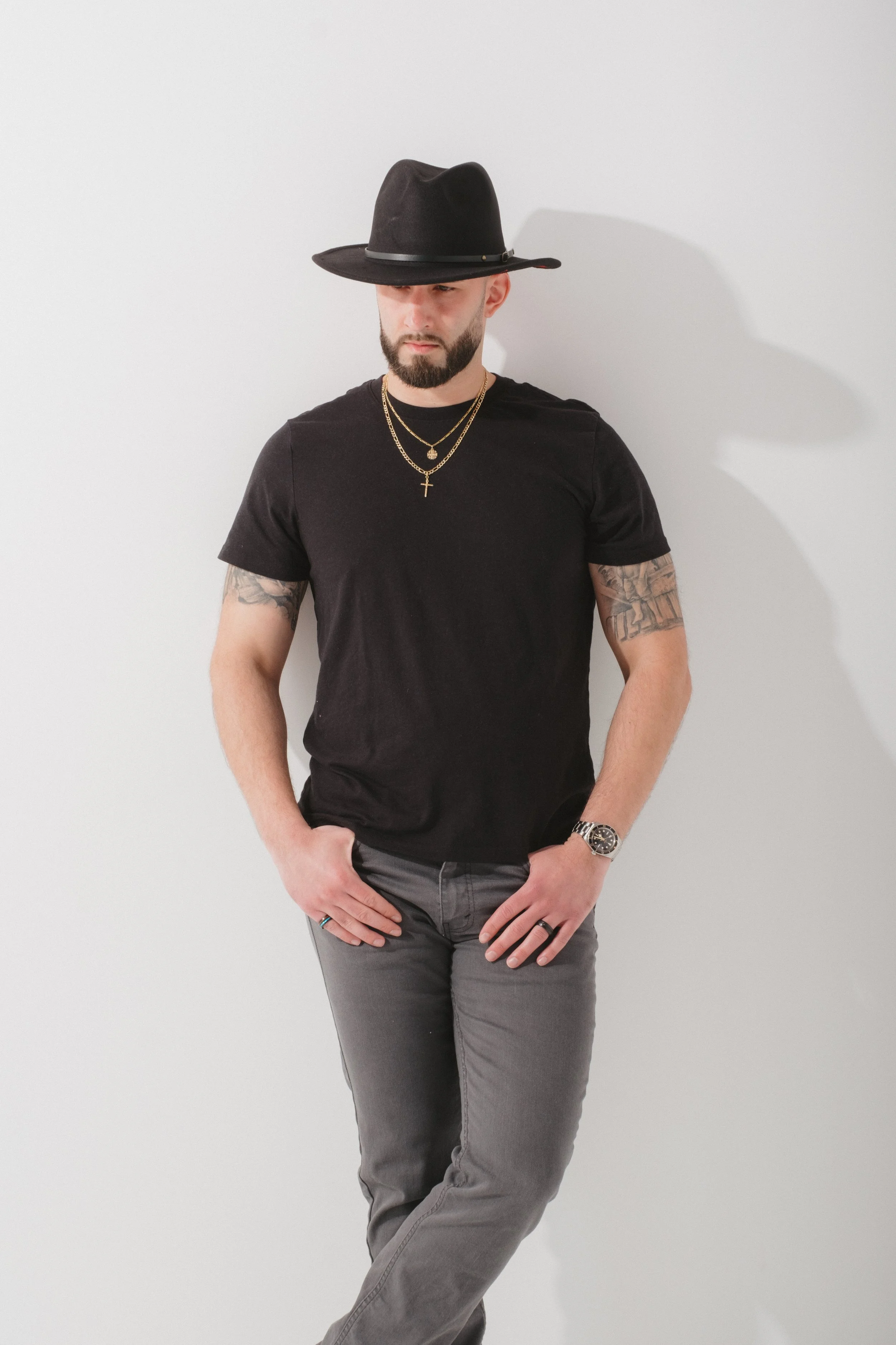 A man with a beard wearing a black hat, black T-shirt, gray pants, gold necklaces, a watch, and rings, standing against a plain white wall.