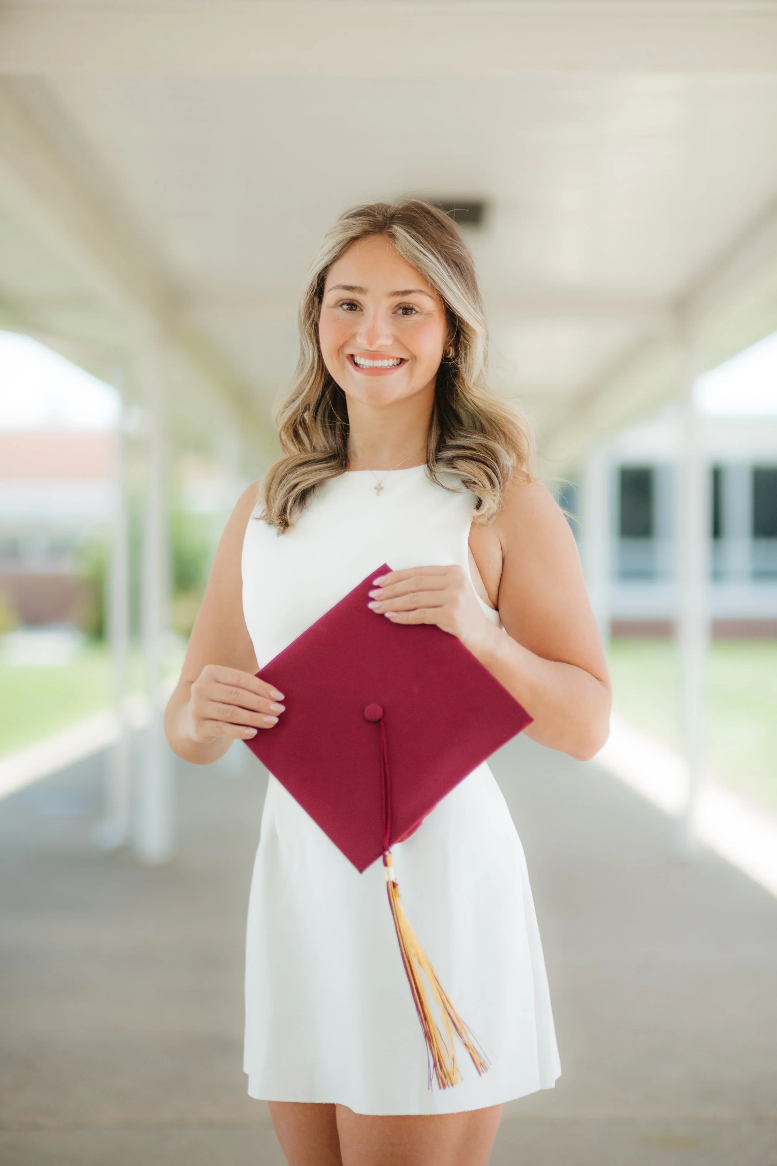 A young woman in a white dress is holding a maroon graduation cap with a gold tassel, smiling in a covered outdoor area.