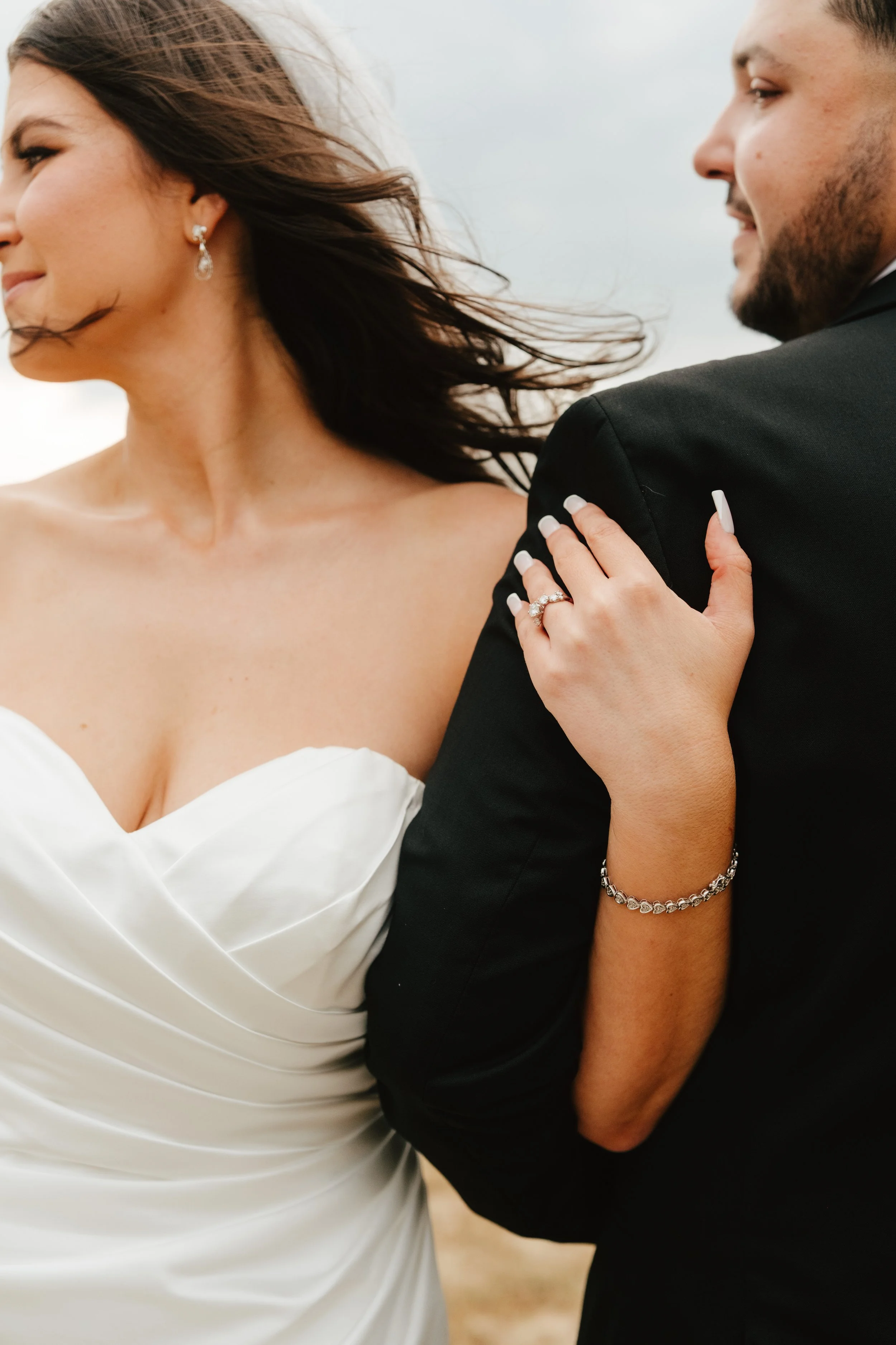 A bride and groom standing close together, holding each other outdoors with wind-blown hair, smiling and showing rings.