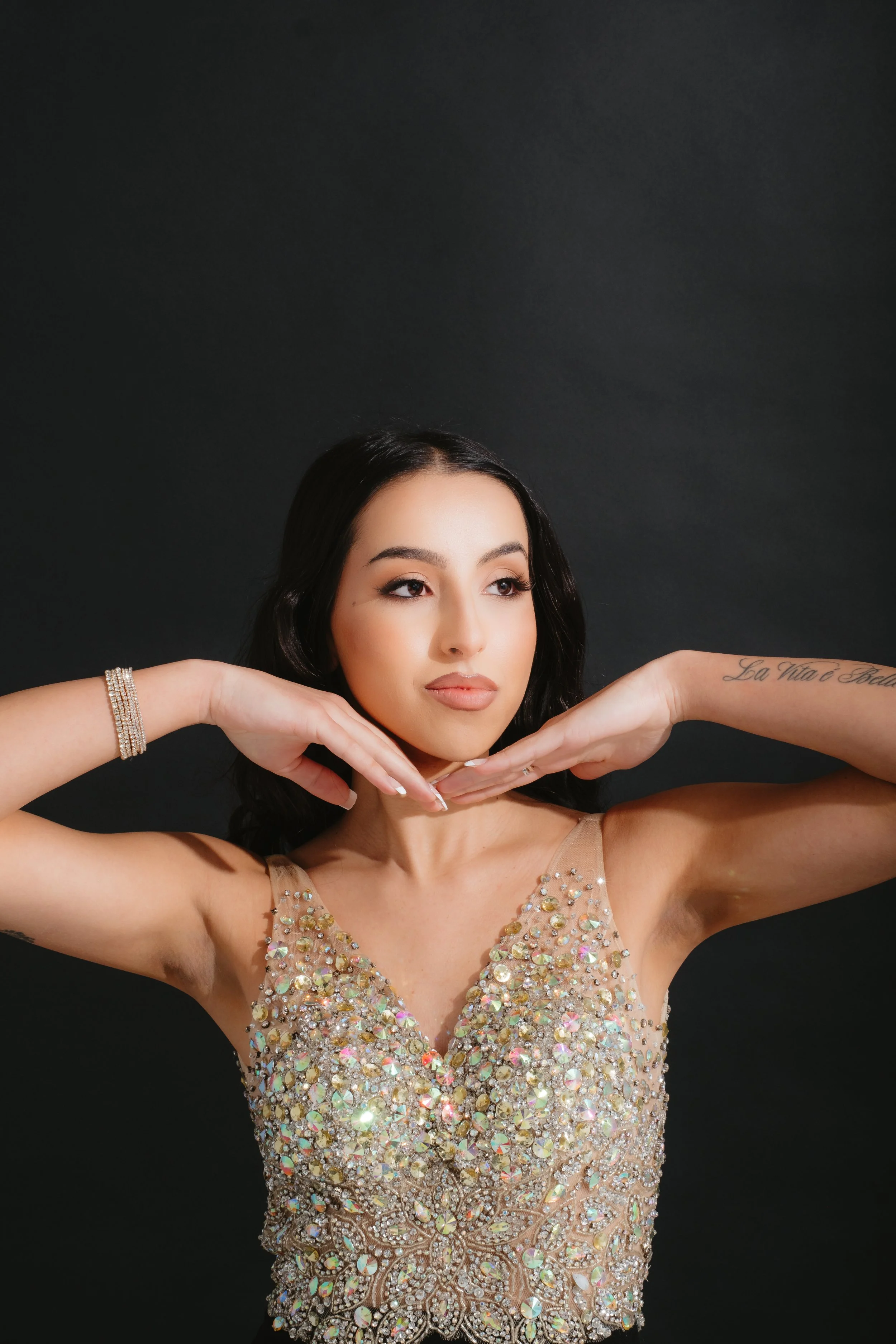 A woman with dark hair, makeup, and a tattoo on her left arm is posing with her hands under her chin, wearing a sleeveless, sparkly, embellished beige dress against a dark background.