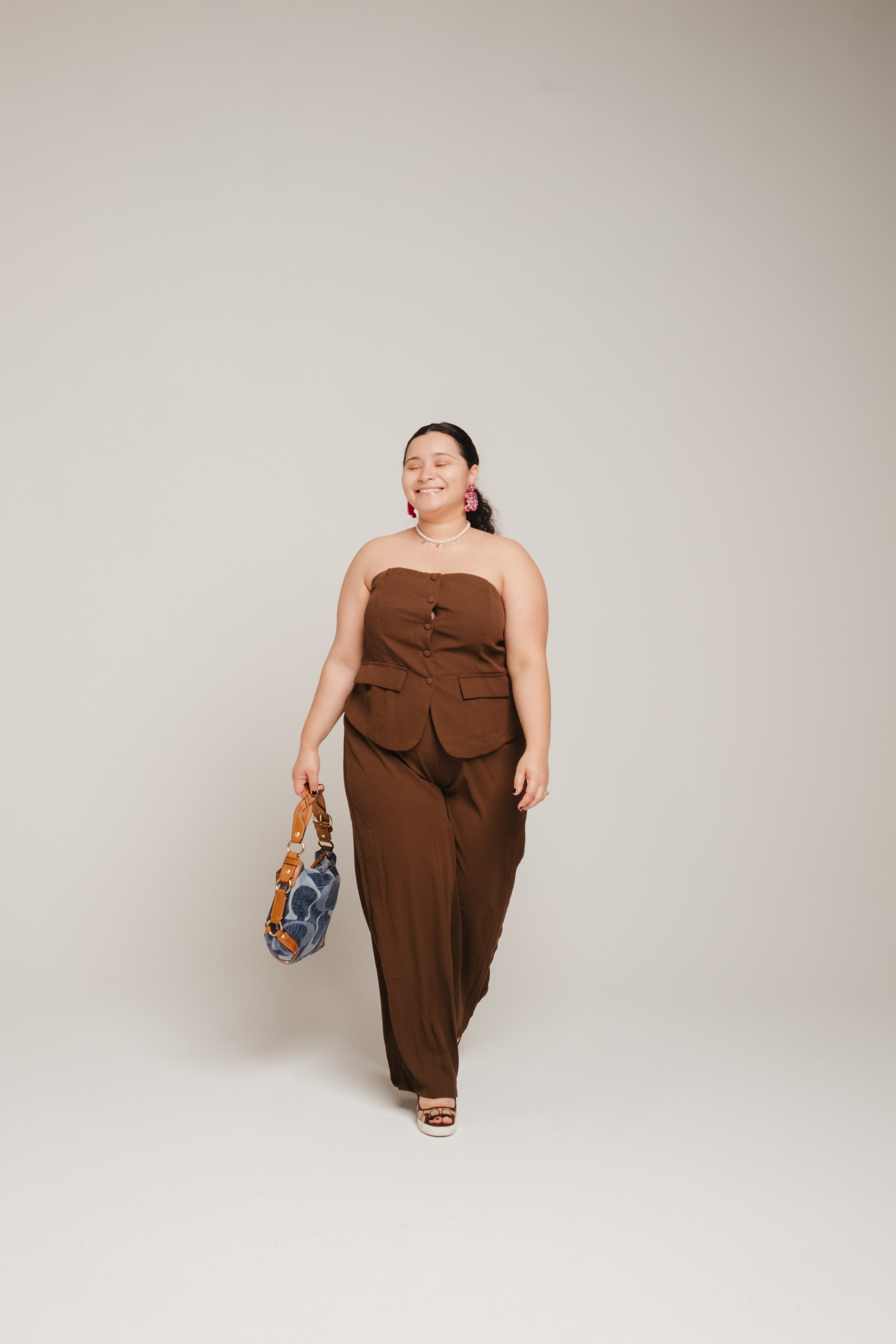 A woman smiling and walking against a plain background, carrying a designer handbag, wearing a brown strapless jumpsuit, sandals, and pink earrings.