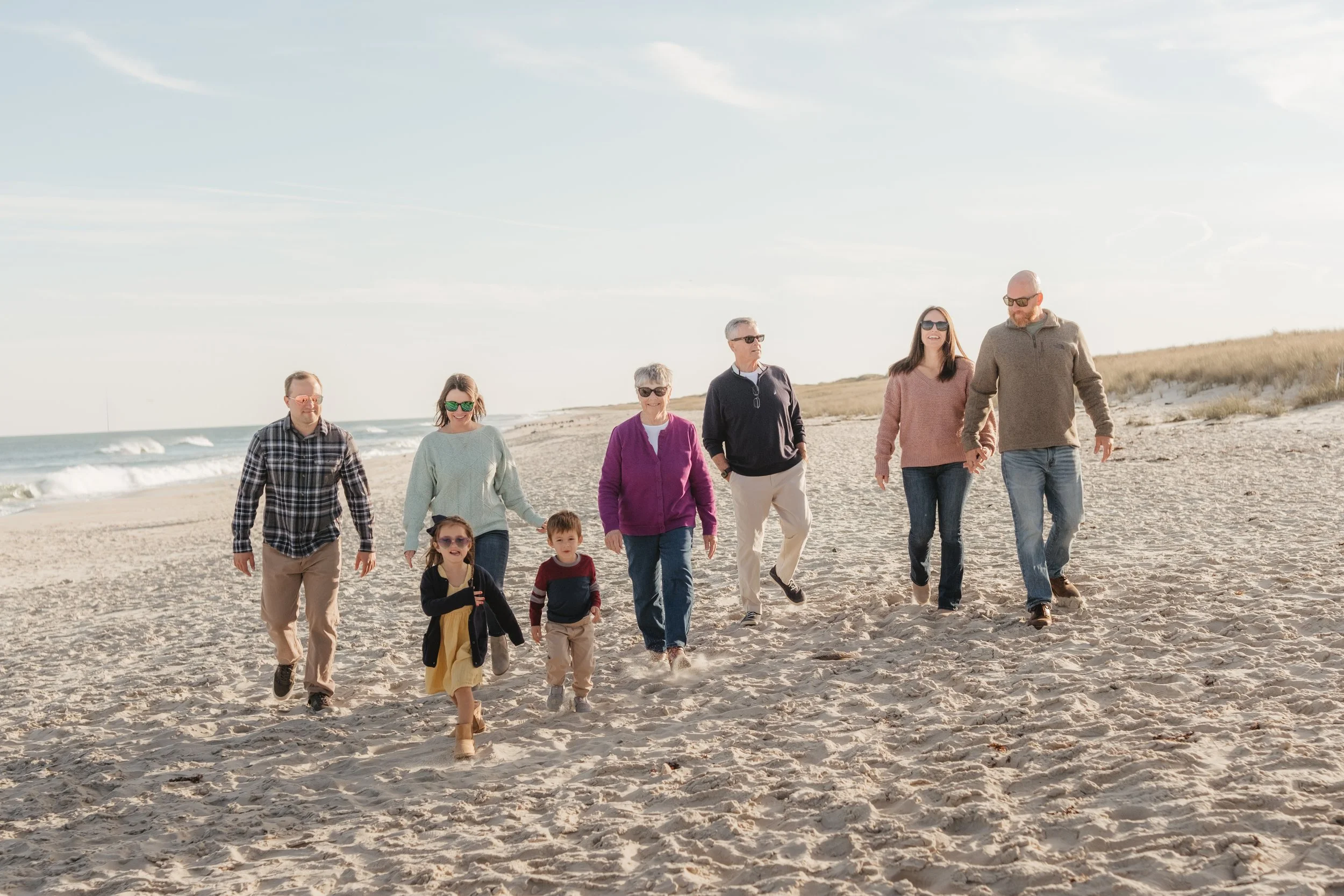 Family walking on the beach with children and adults holding hands. The beach has sand dunes and the ocean in the background. It is a sunny day.