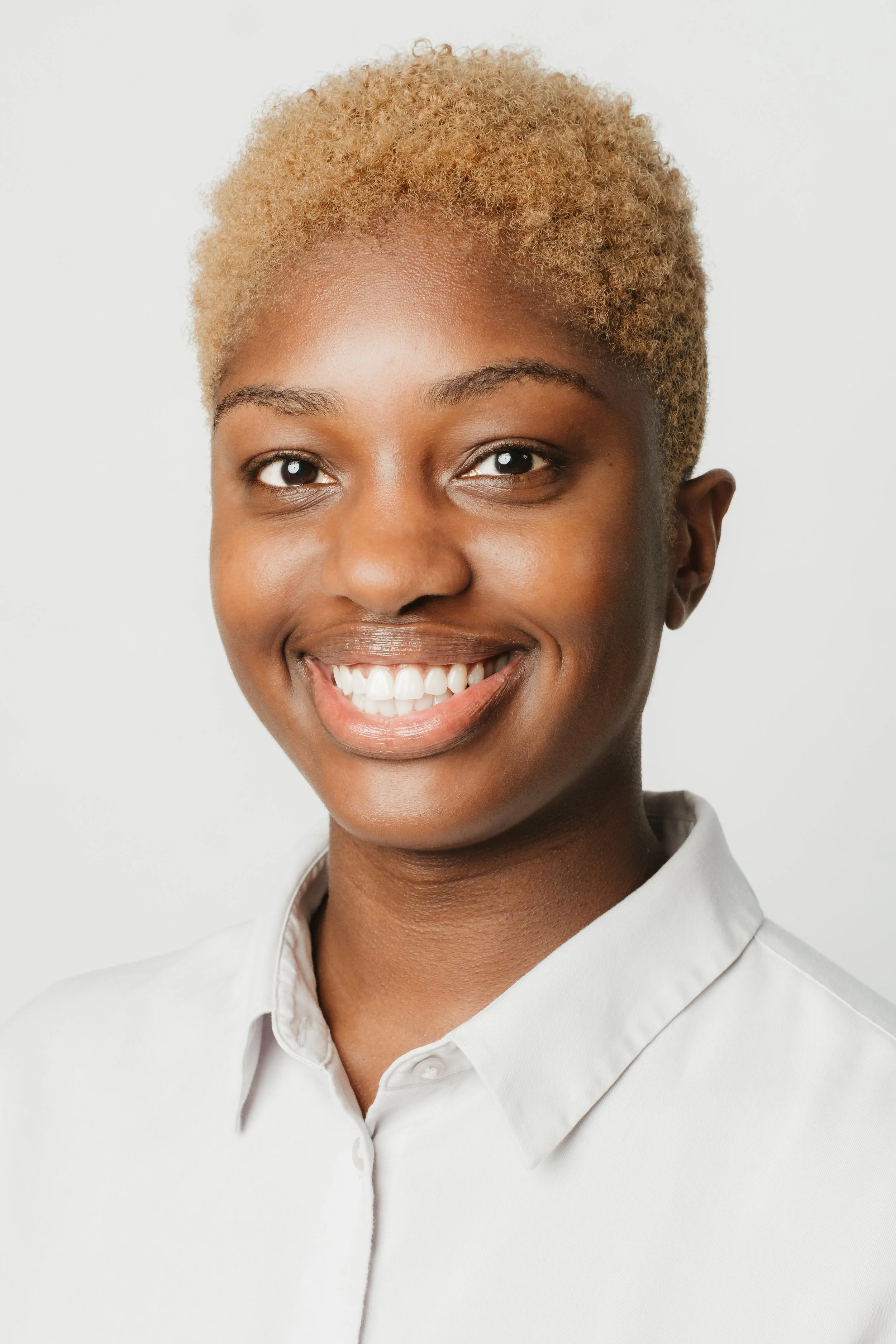 Close-up of a smiling woman with short, curly blonde hair wearing a white collared shirt against a plain white background.