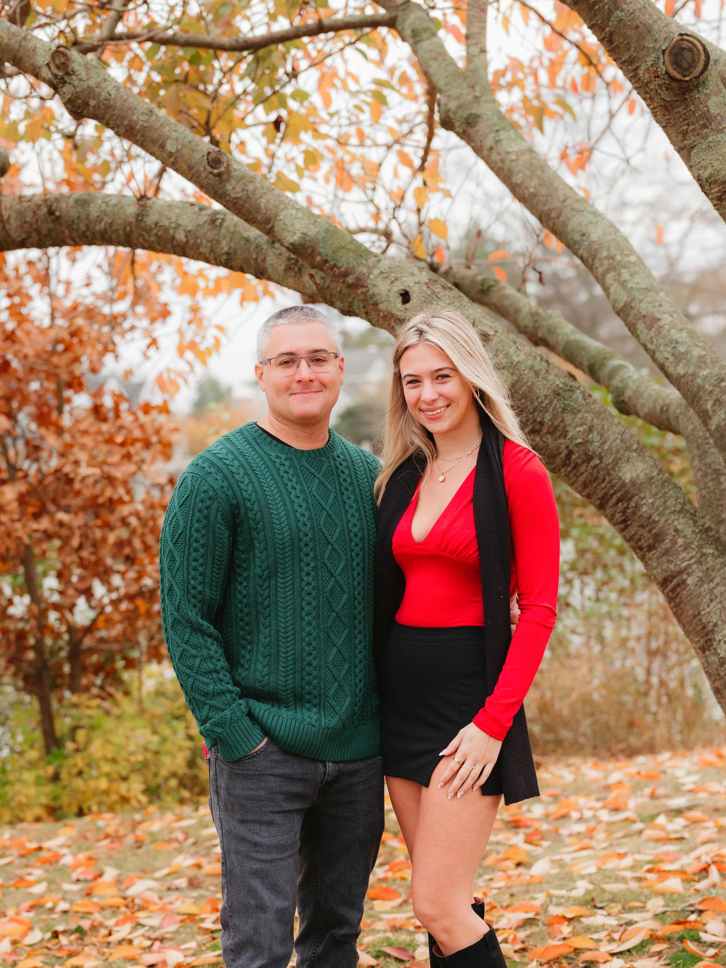 A man and woman standing outdoors during autumn, with colorful fall leaves on trees and ground, smiling at the camera.