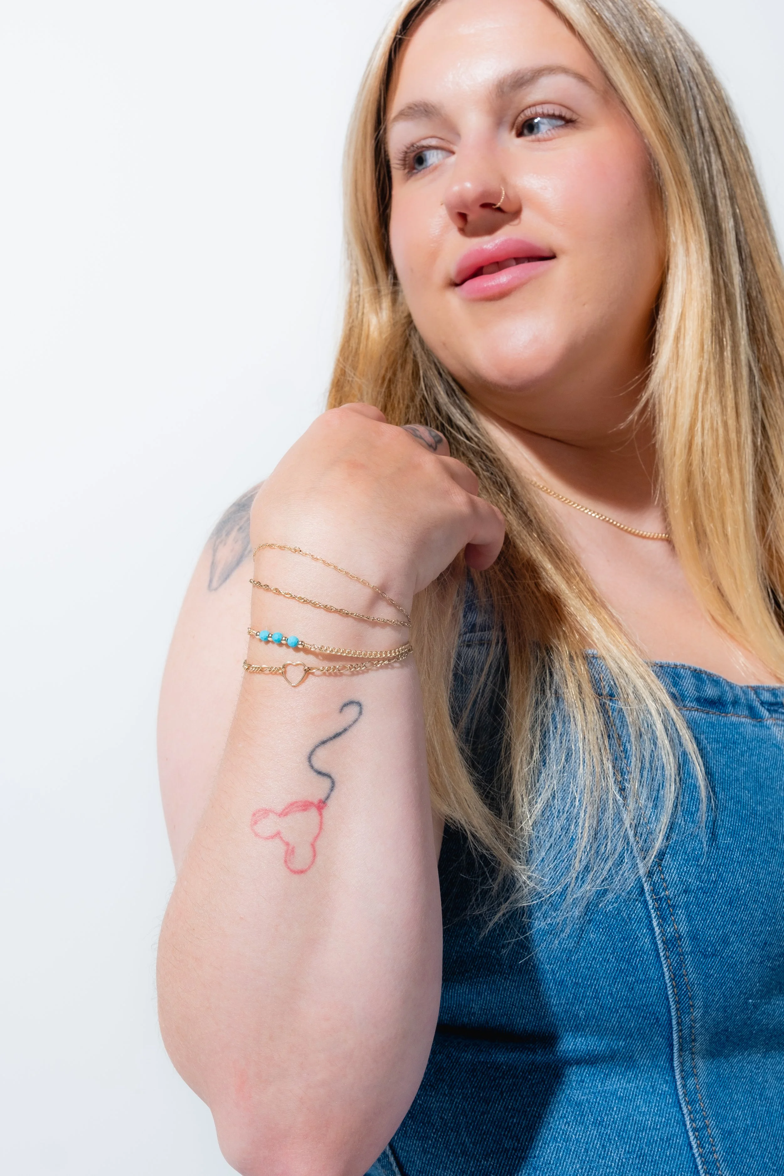 A woman with blonde hair, wearing a denim top, showing colorful bracelets and a tattoo of a squiggly line and a red heart on her forearm.
