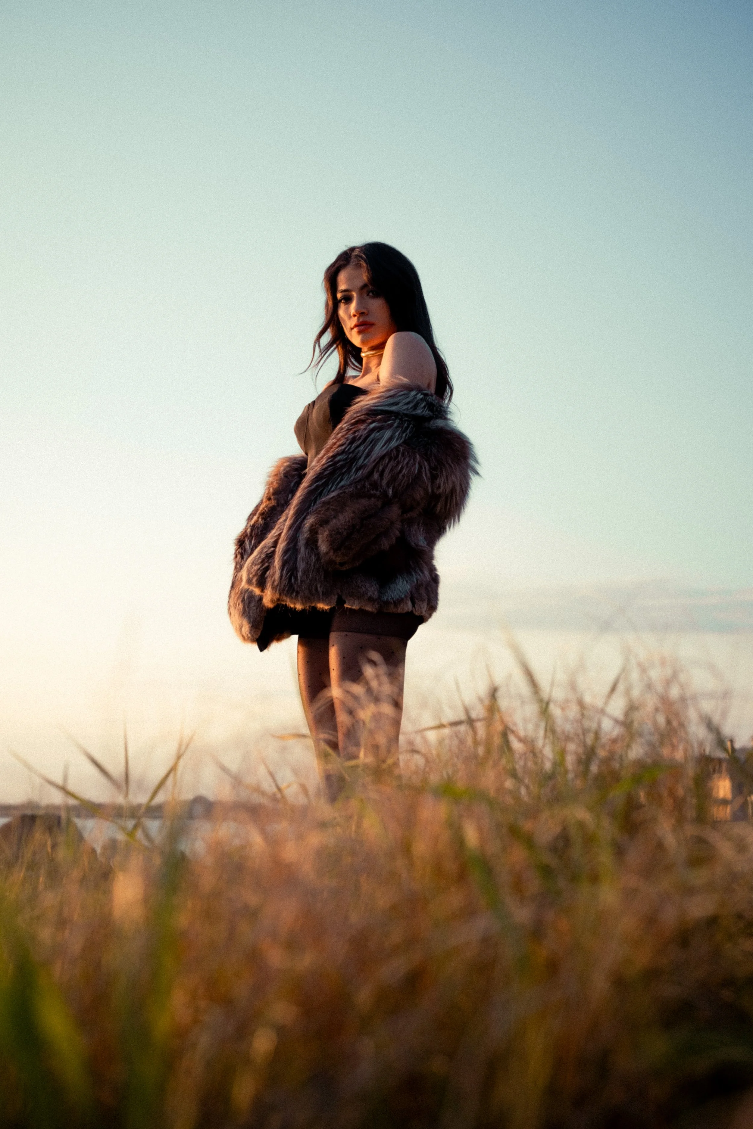 A woman standing in a field at sunset, wearing a fur coat over a black dress and tights.