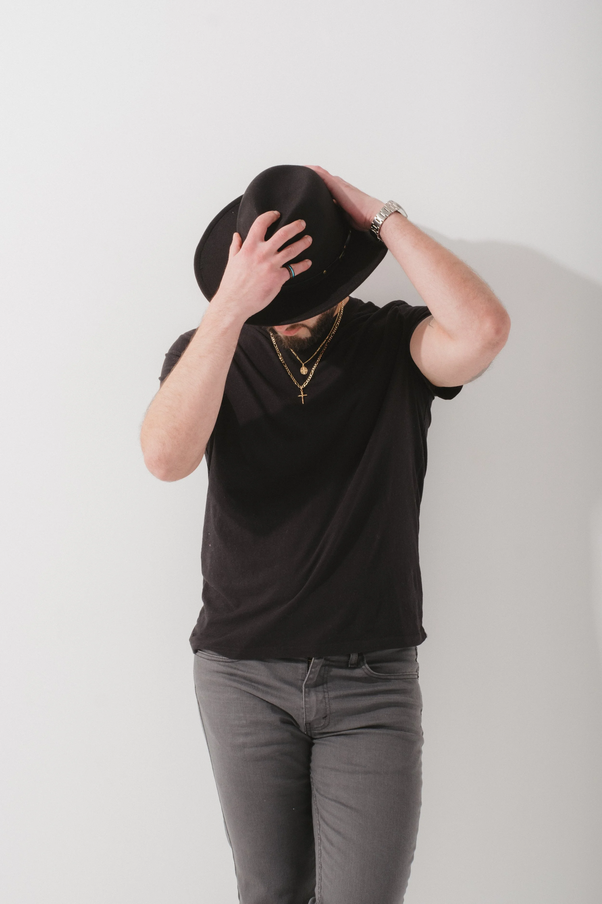 Person wearing a black t-shirt and gray jeans holding a black hat over their face, standing against a plain white background.