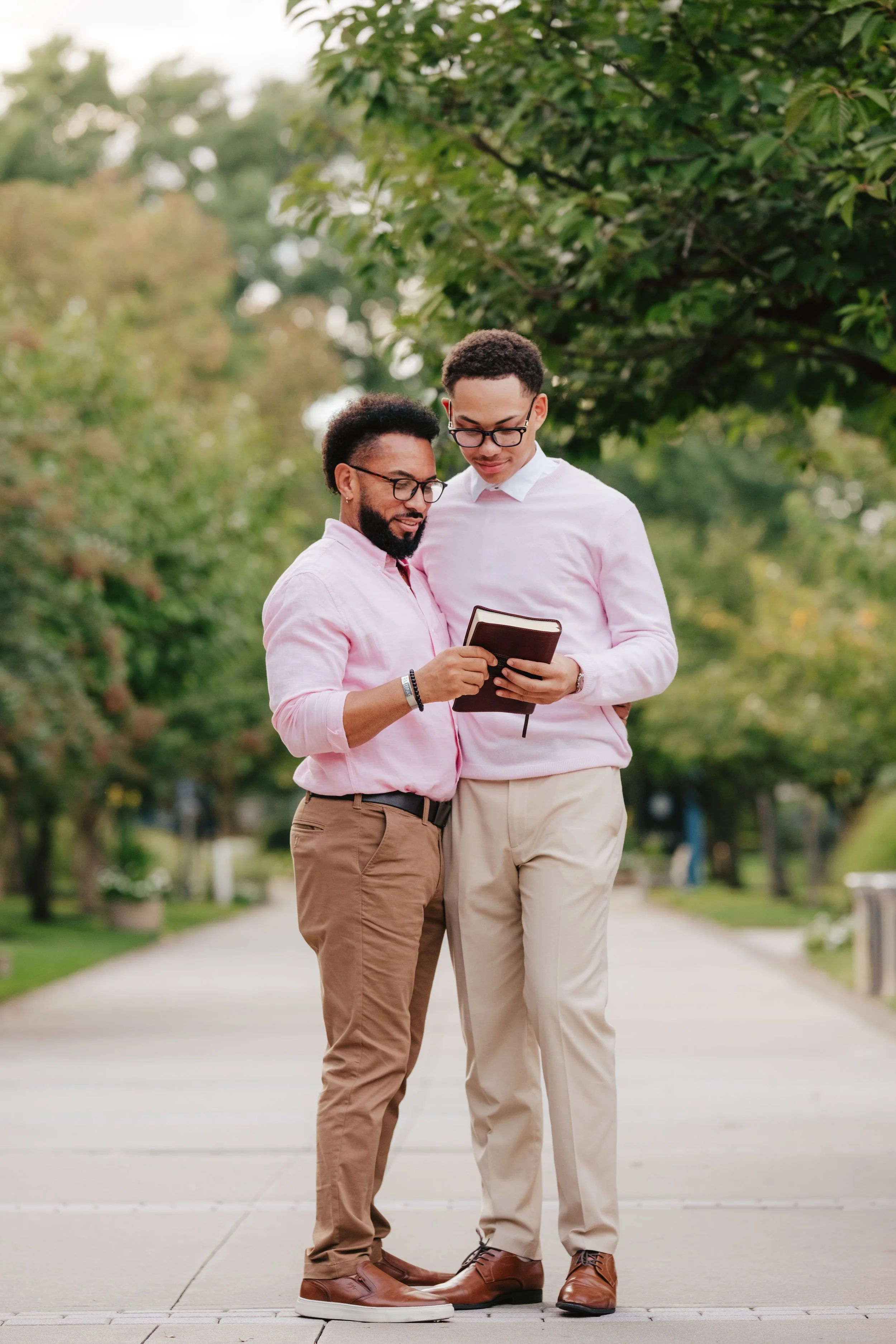 Two men standing outdoors on a paved walkway, looking at a book together, surrounded by green trees.
